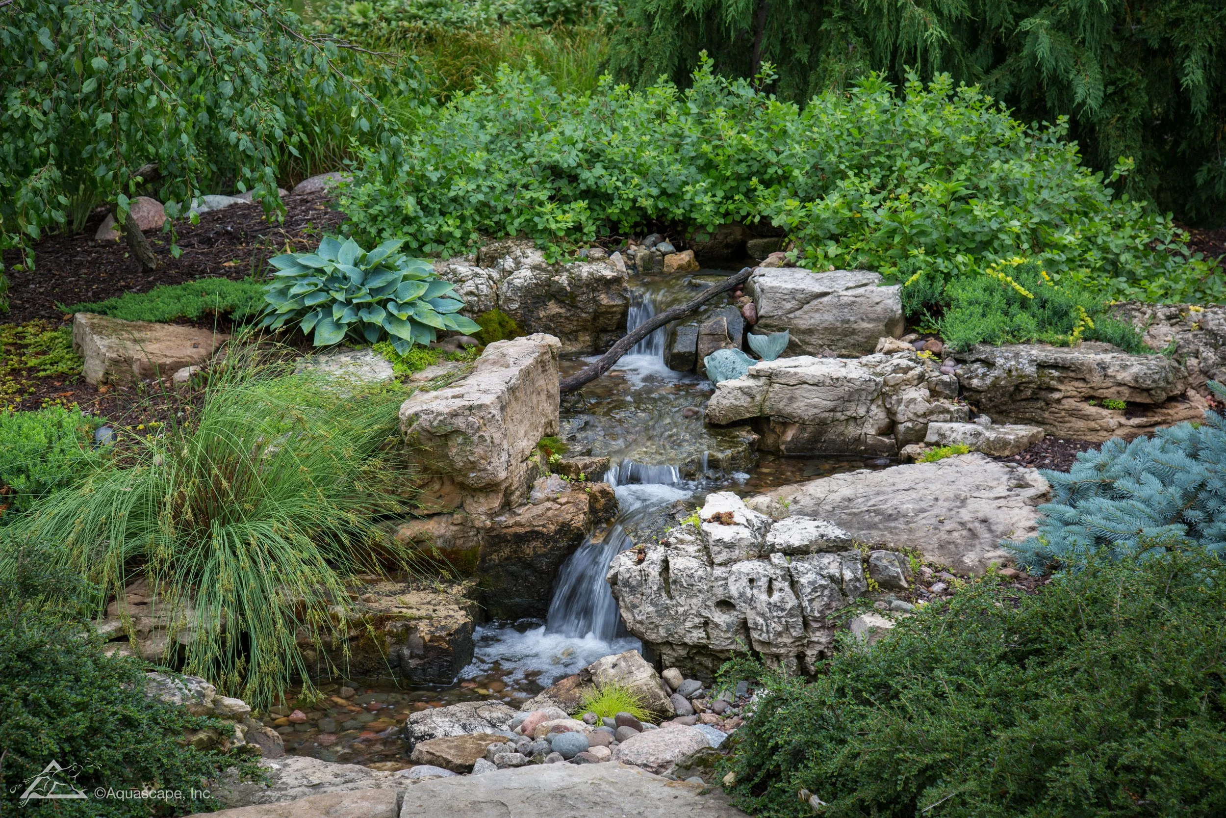 A small waterfall flowing over rocks in a green, lush garden with various plants and bushes.