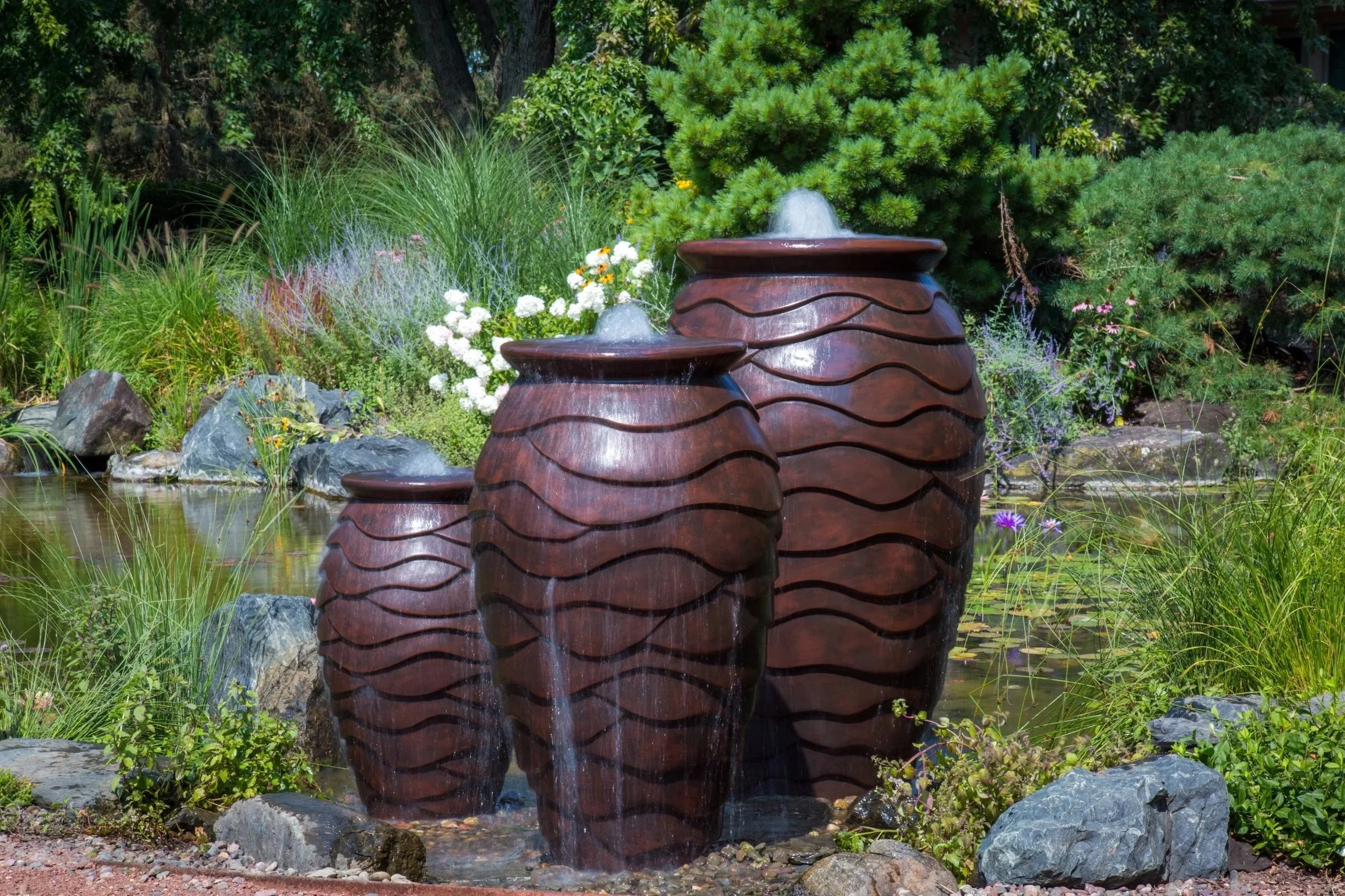Three brown ceramic garden fountains with water flowing from the top, surrounded by green plants and rocks in a garden setting.