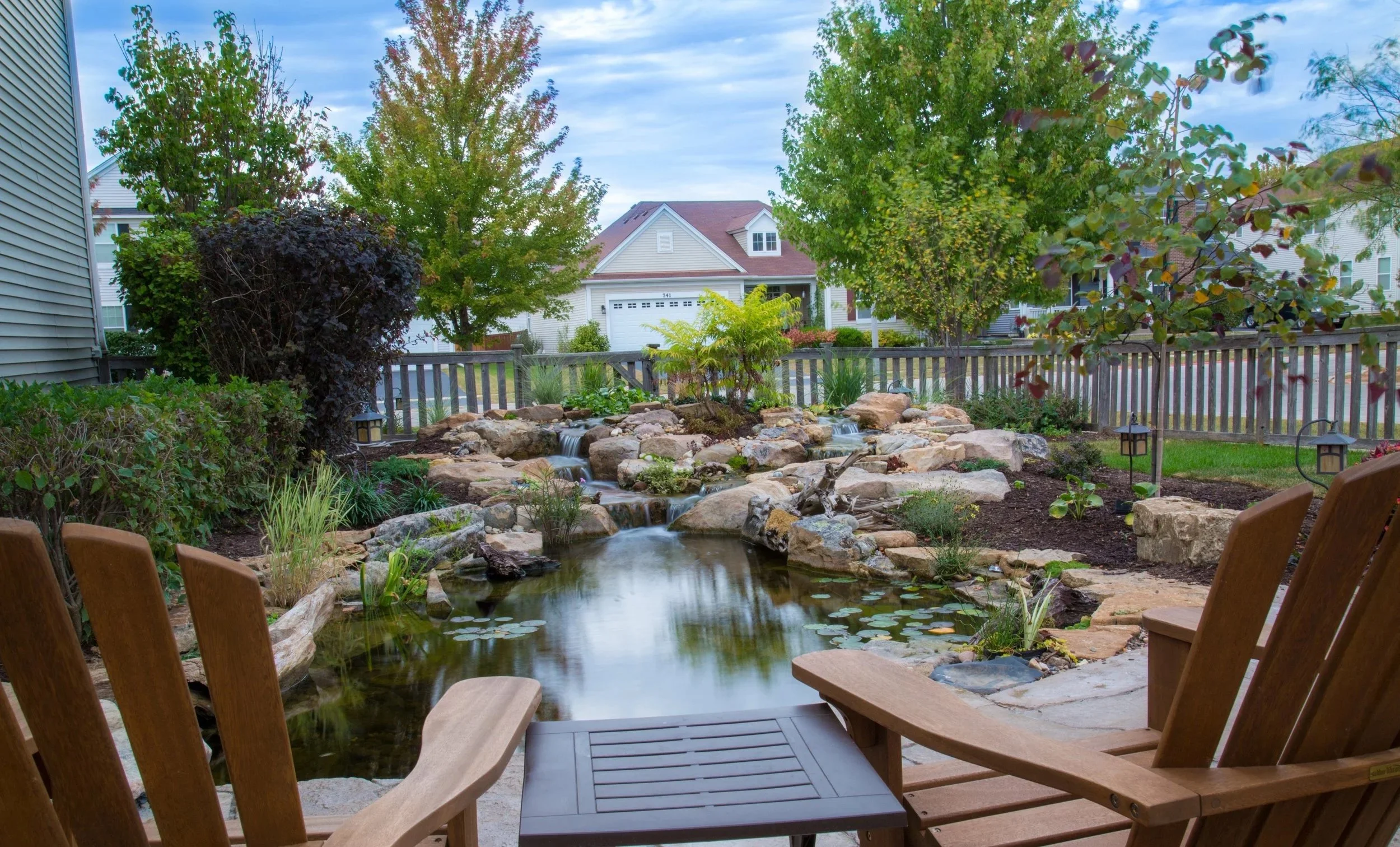 A backyard with a pond, small waterfall, and garden surrounded by trees, with neighboring houses visible in the background.