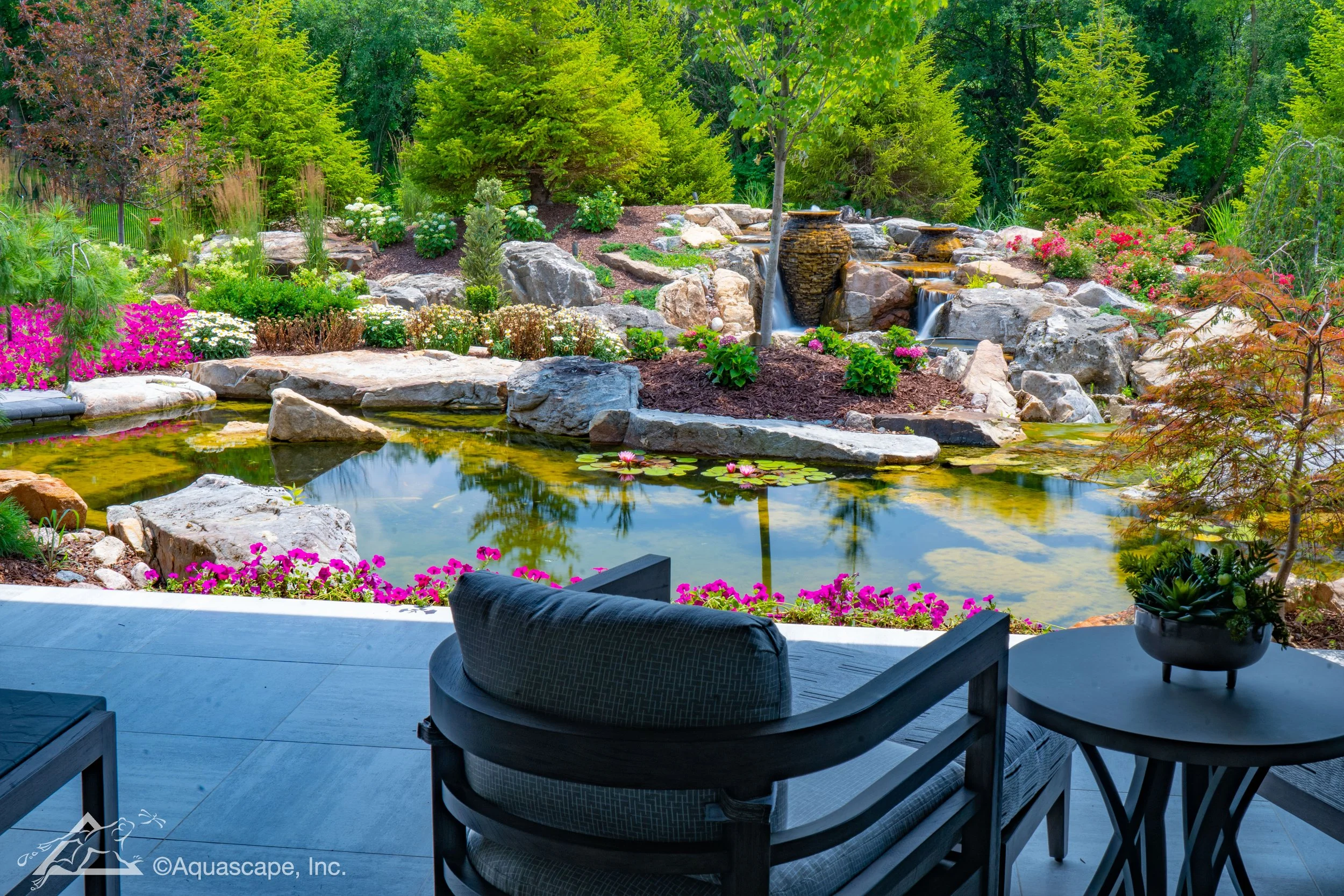 View from a patio overlooking a landscaped garden with a pond, rocks, green trees, and colorful flowers.
