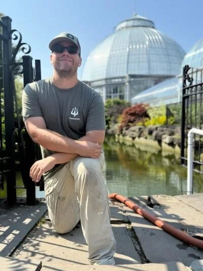 A man kneeling on a wooden dock near a pond, wearing sunglasses, a baseball cap, a gray t-shirt, and khaki pants, with a large glass greenhouse and lush landscaping in the background.
