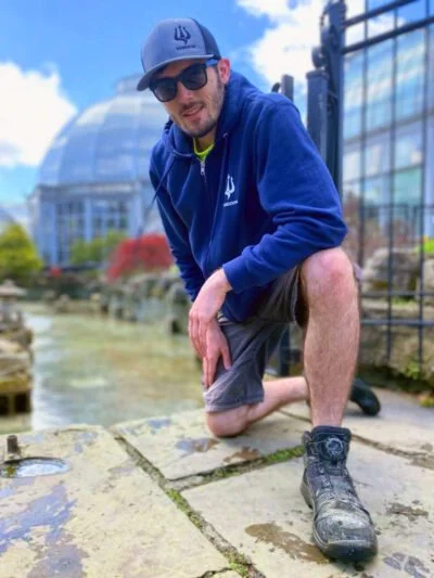 A man wearing sunglasses, a blue hoodie, a matching cap, and gray shorts crouches on a stone pathway outdoors near glass greenhouse structures, with flowering trees and a partly cloudy sky in the background.