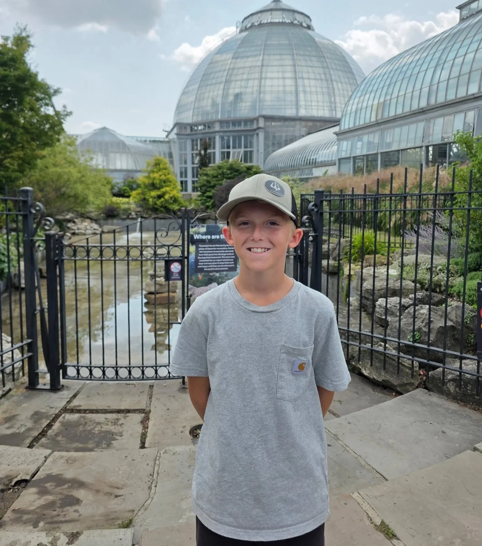 Young boy smiling in front of a greenhouse in a botanical garden.