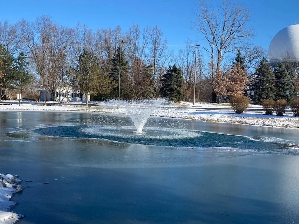 A frozen pond with a small fountain spray in the center, surrounded by snow and bare trees, under a clear blue sky.