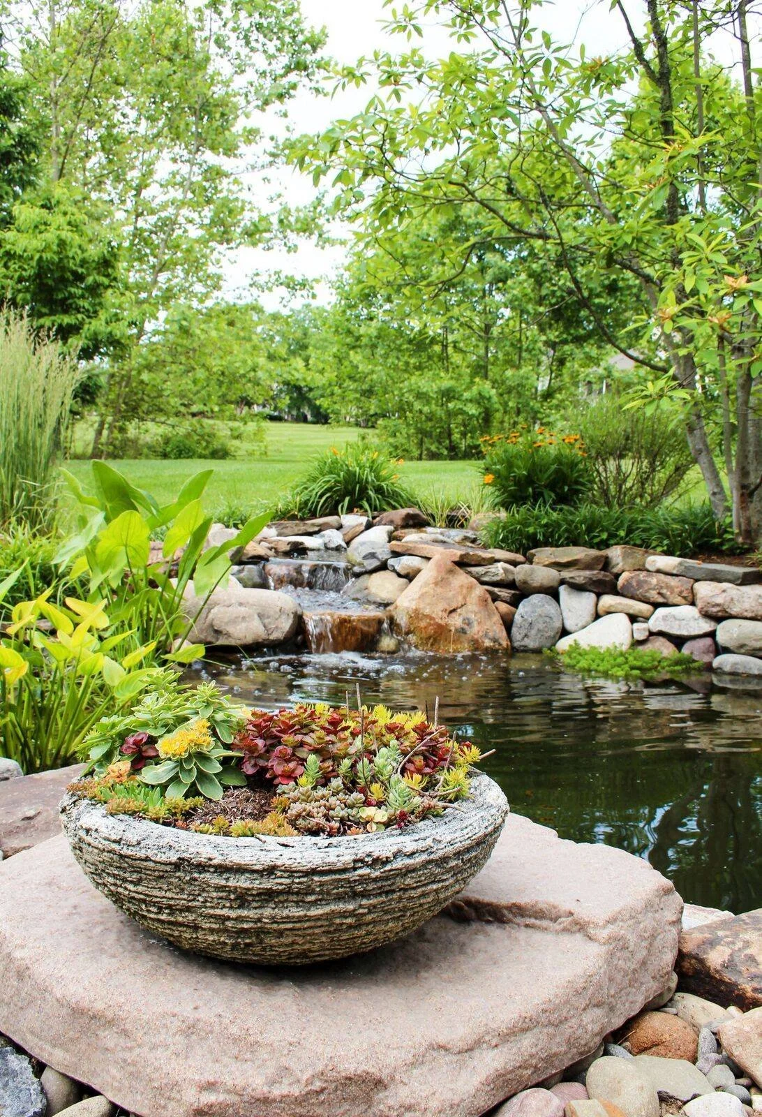 A garden with a small waterfall, surrounded by rocks, lush green plants, and trees, with a grassy lawn in the background.
