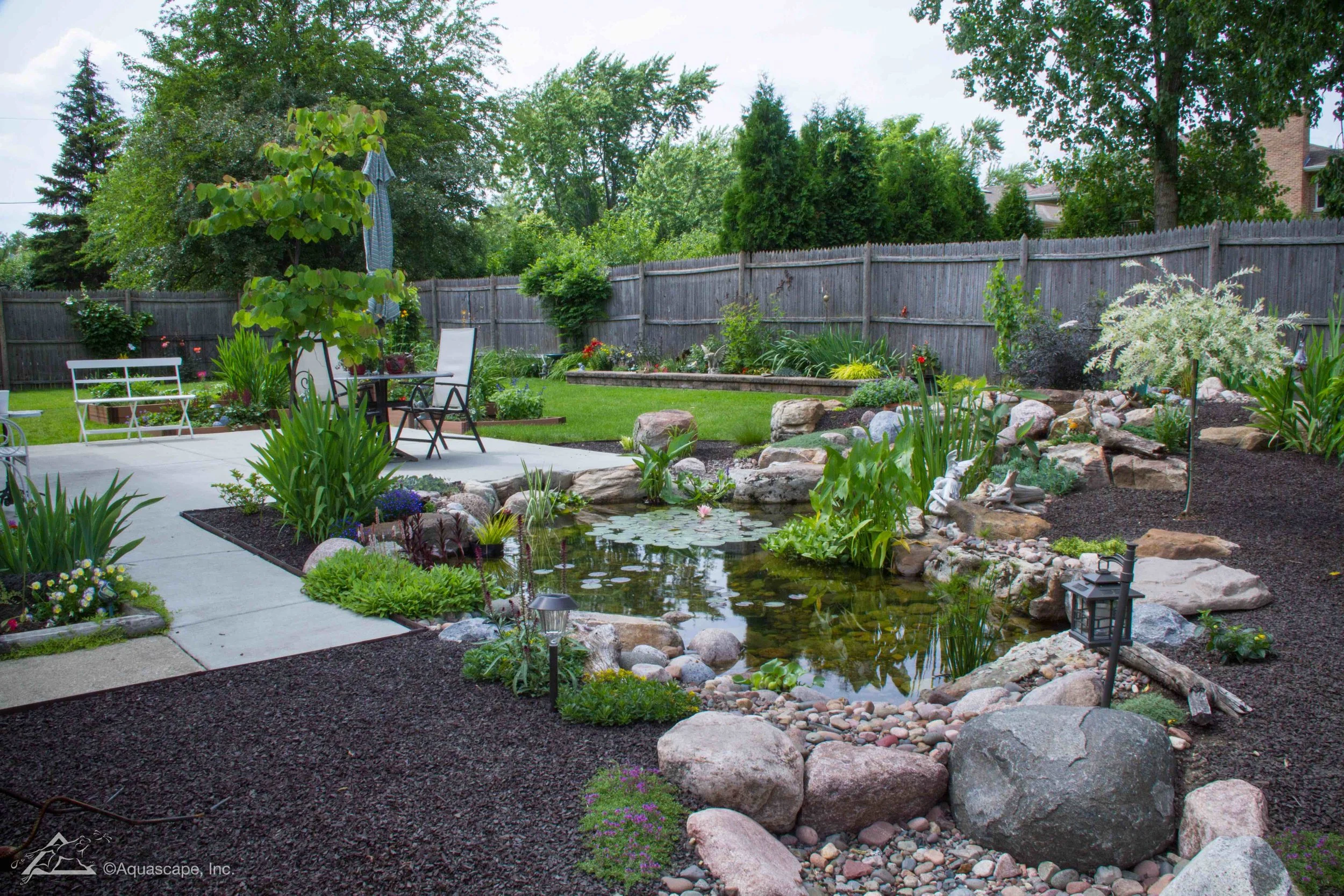 A backyard with a pond surrounded by rocks, green plants, and flowers, a patio with a table and chairs, a wooden fence, and trees in the background.