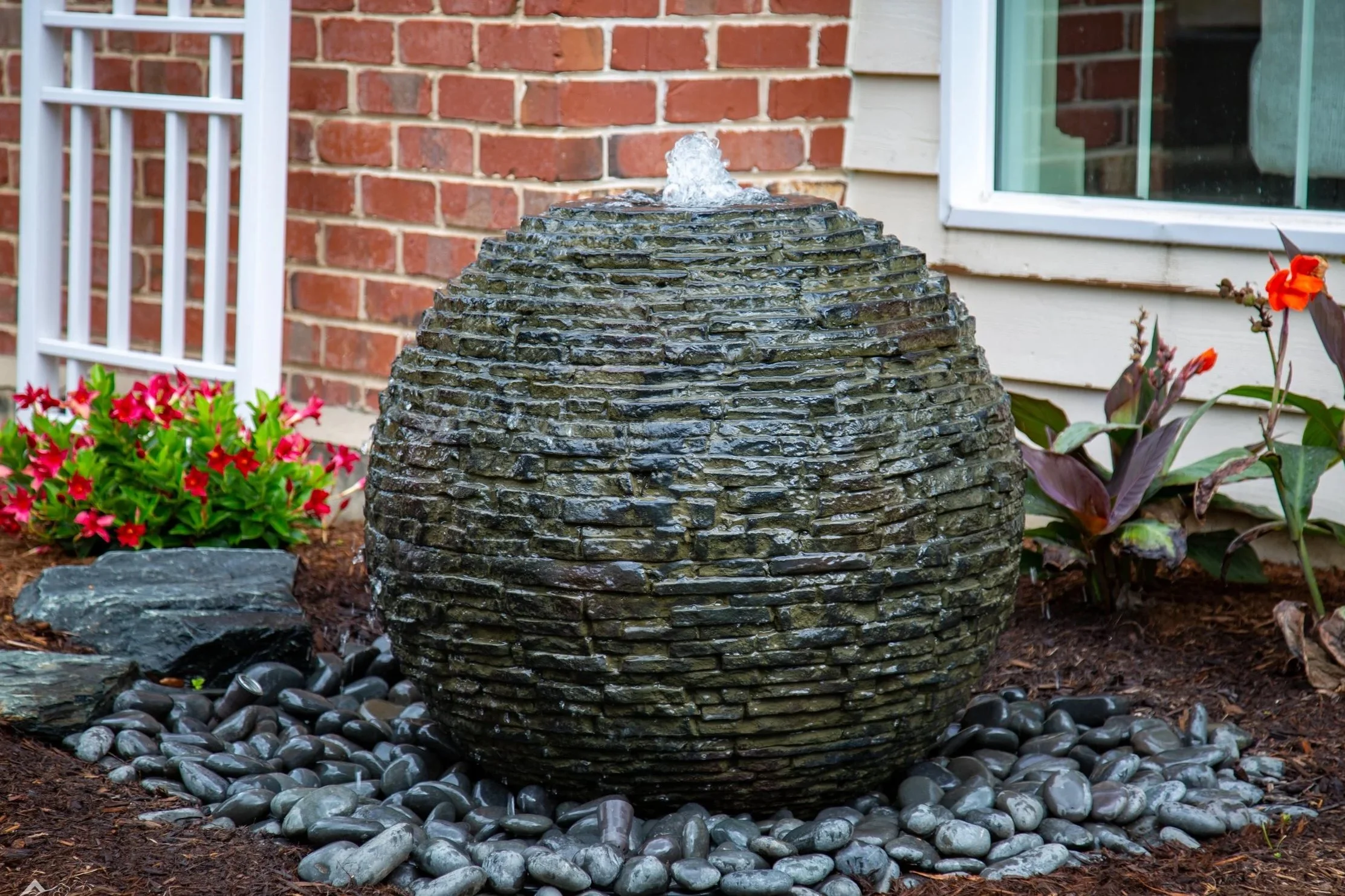 A small landscaped garden with a water fountain made of stacked flat stones, surrounded by black pebbles and colorful flowers, next to a brick house wall and a window.