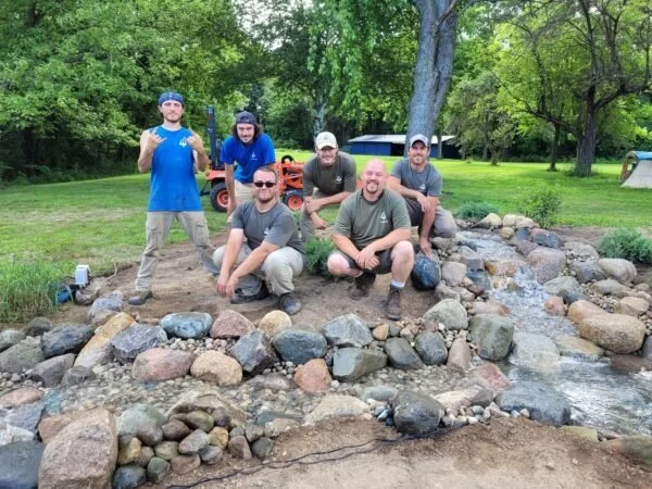 Six men posing by a small backyard stream surrounded by rocks, with trees in the background.