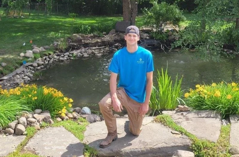 Young man kneeling on a stone pathway beside a pond with yellow flowers, green plants, and trees in the background.
