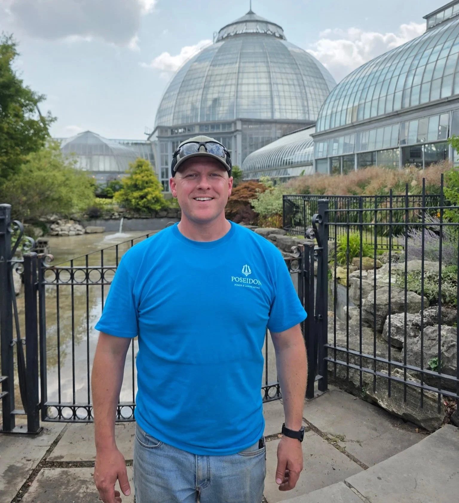 A man smiling outdoors in front of a glass-domed greenhouse with lush green plants and a small pond, wearing a blue t-shirt, sunglasses on his cap, and a watch.