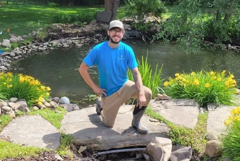 Man with glasses, beard, and tattoos kneeling on one knee outdoors by a pond, surrounded by yellow flowers and green foliage.