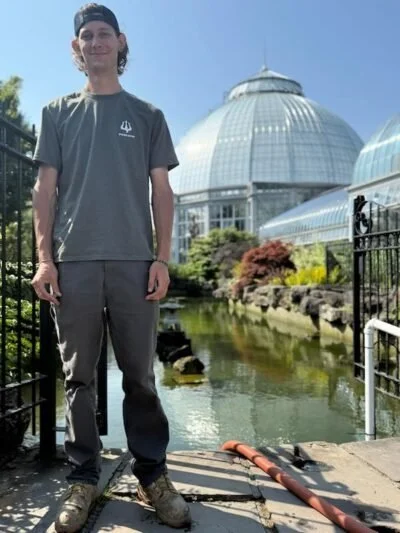 A young man standing outdoors near a pond with a greenhouse in the background.