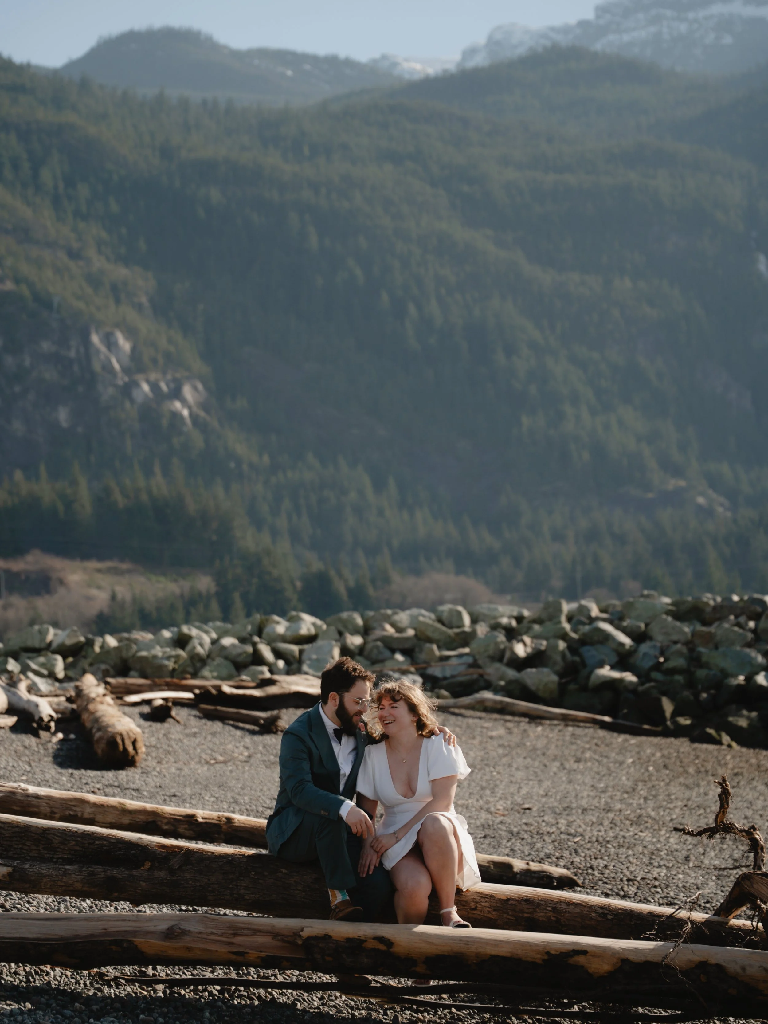 A couple sitting on driftwood logs on a rocky beach with mountains and a forest in the background, smiling and enjoying each other's company.