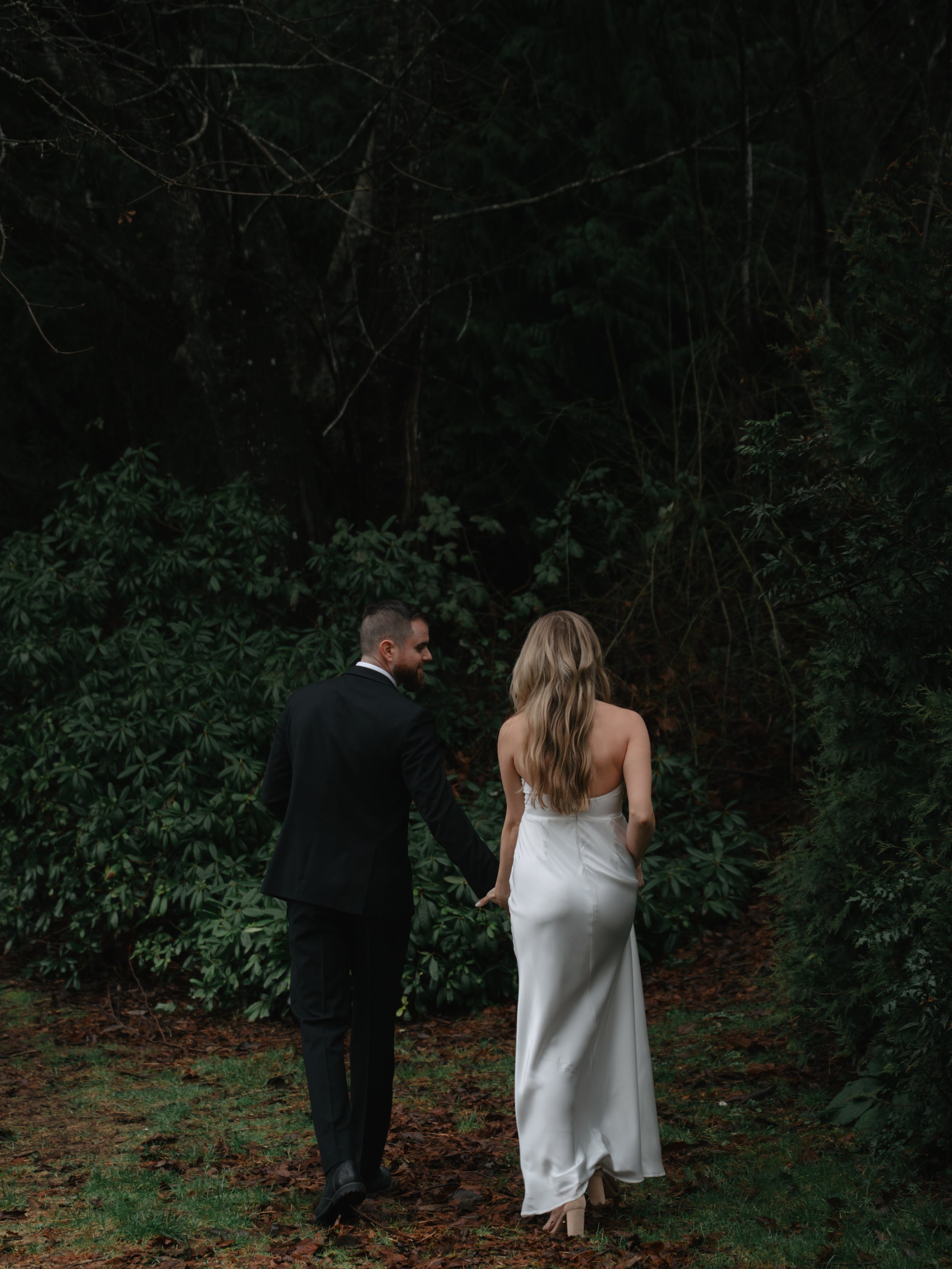 A couple dressed in wedding attire holding hands and walking through a forested area.