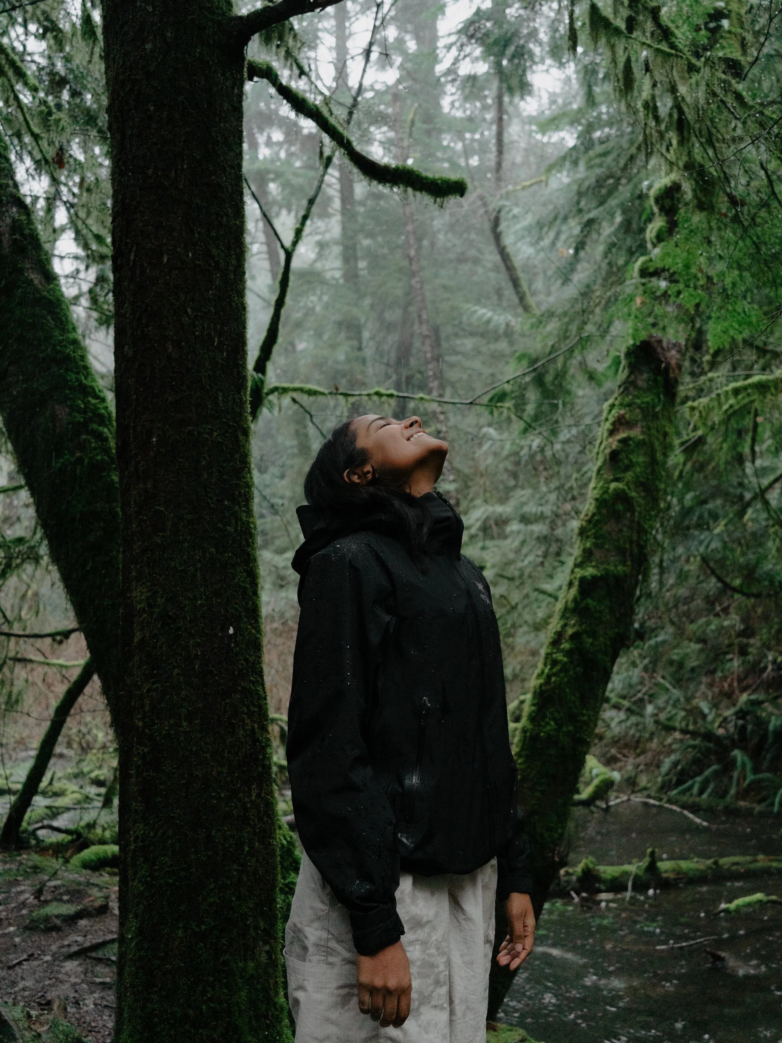 A woman in a black rain jacket and beige pants stands in a foggy, mossy forest, smiling and looking up at the trees.