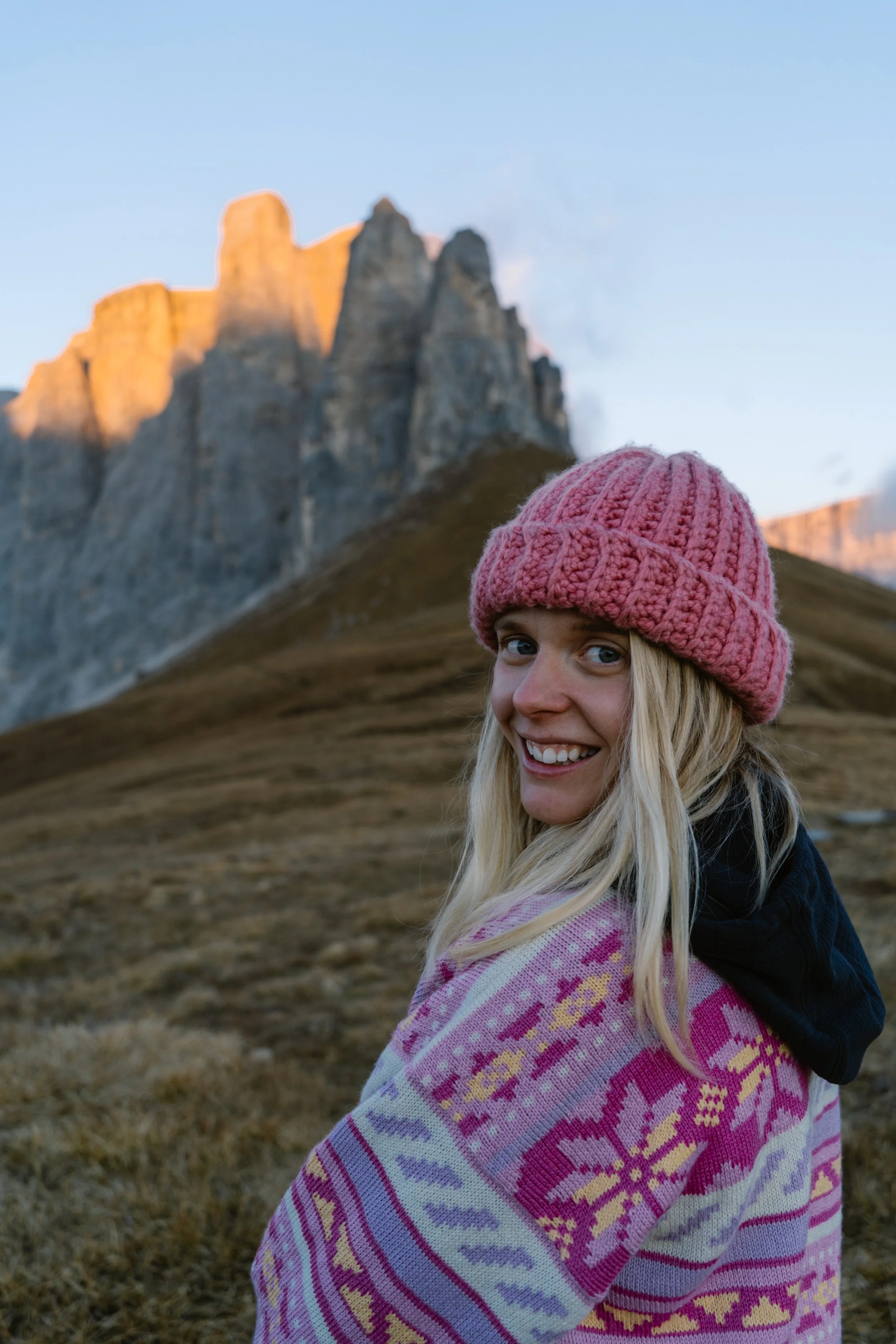 A smiling blonde woman wearing a pink knit hat and a colorful patterned shawl, standing outdoors with mountains in the background during sunset.