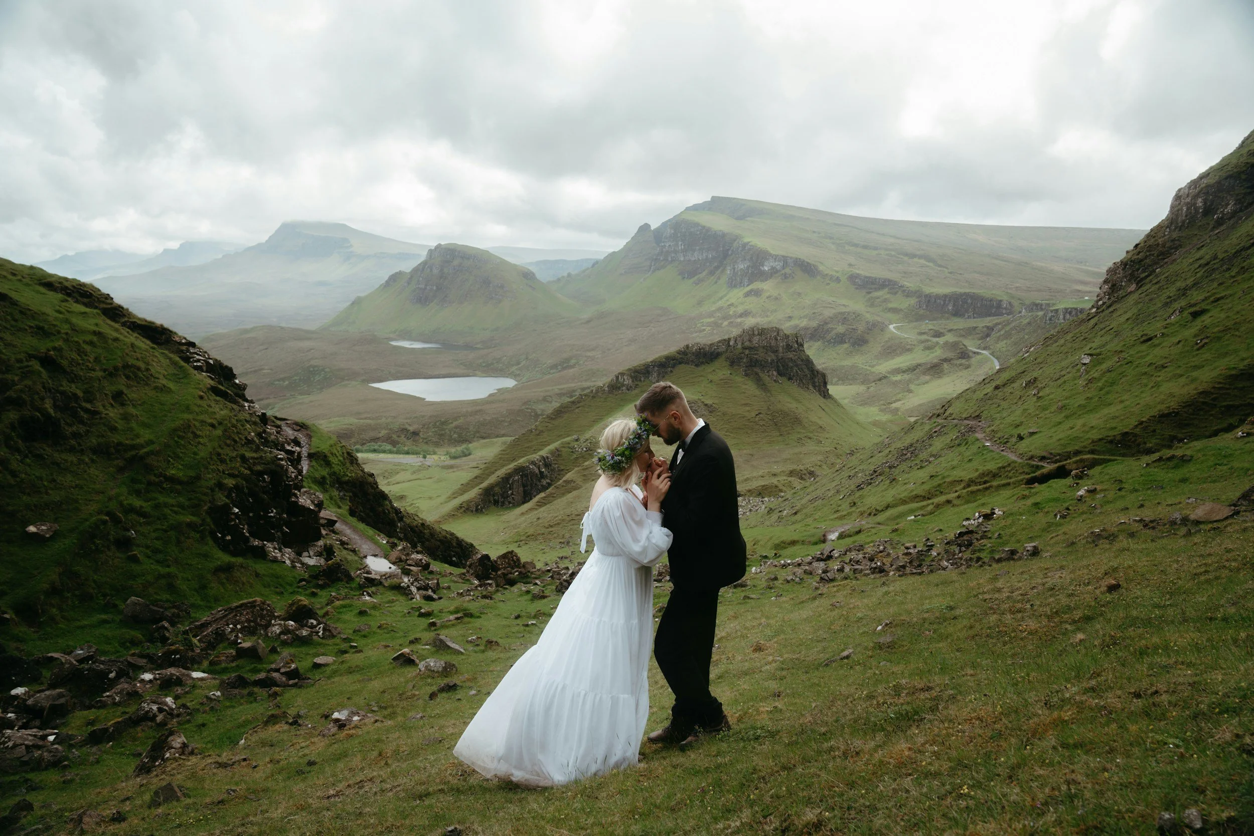 A bride and groom share a tender moment in a lush green valley with rolling hills and distant lakes, under a cloudy sky.