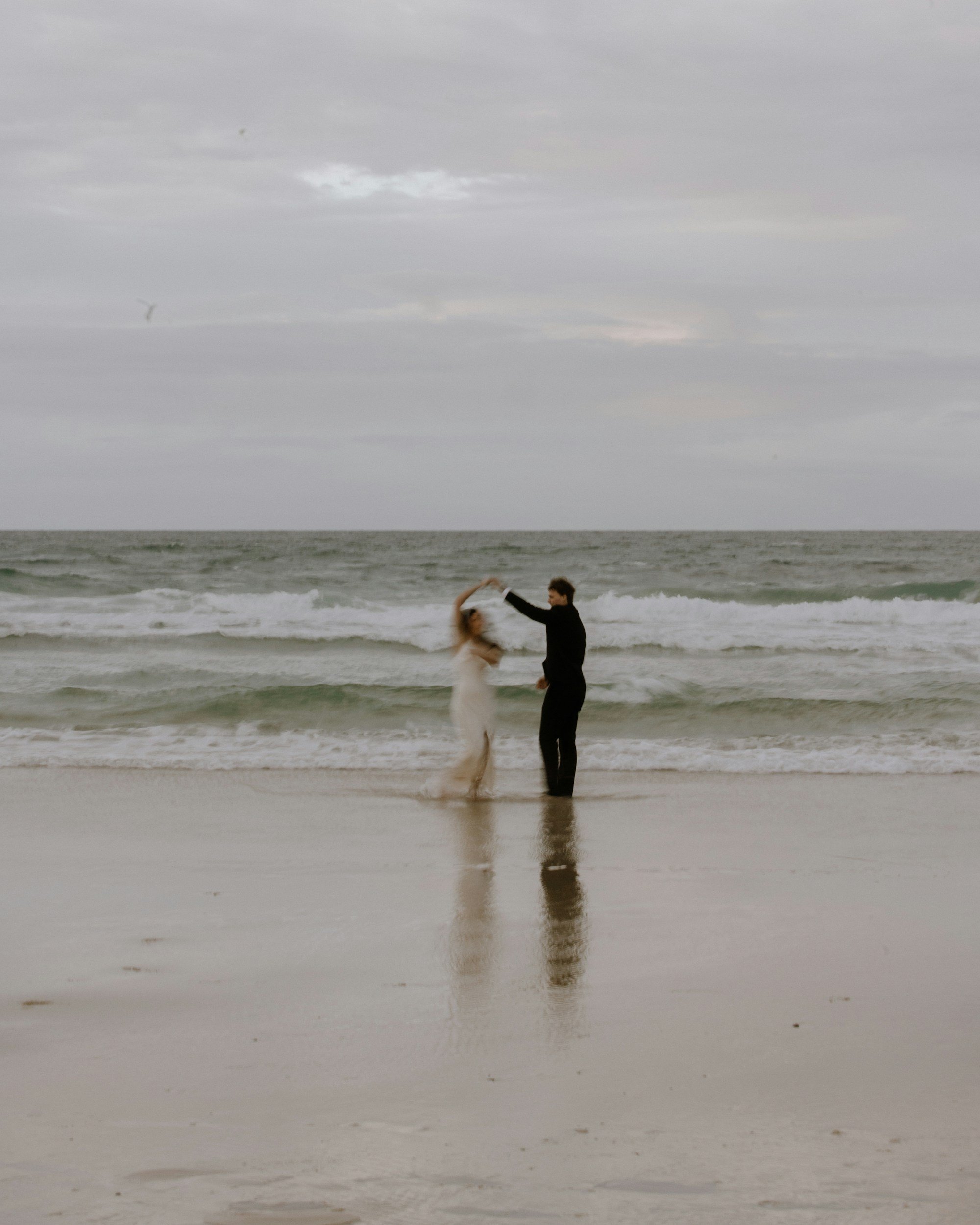 A couple dancing on the beach by the ocean, with cloudy skies overhead.