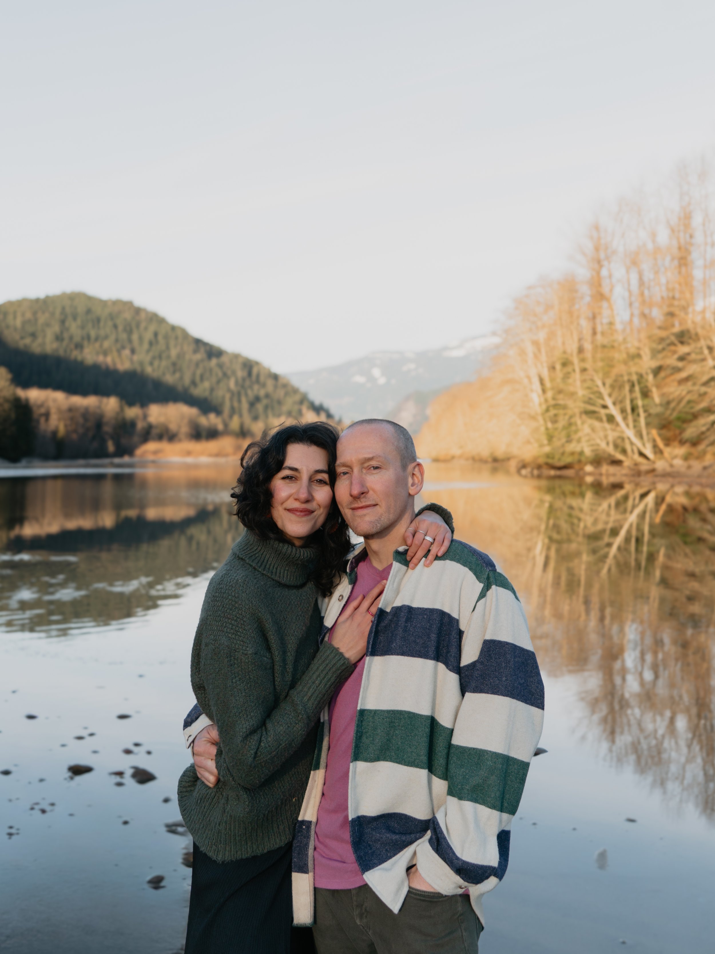 A couple standing by a calm lake with mountains and trees in the background during sunset.