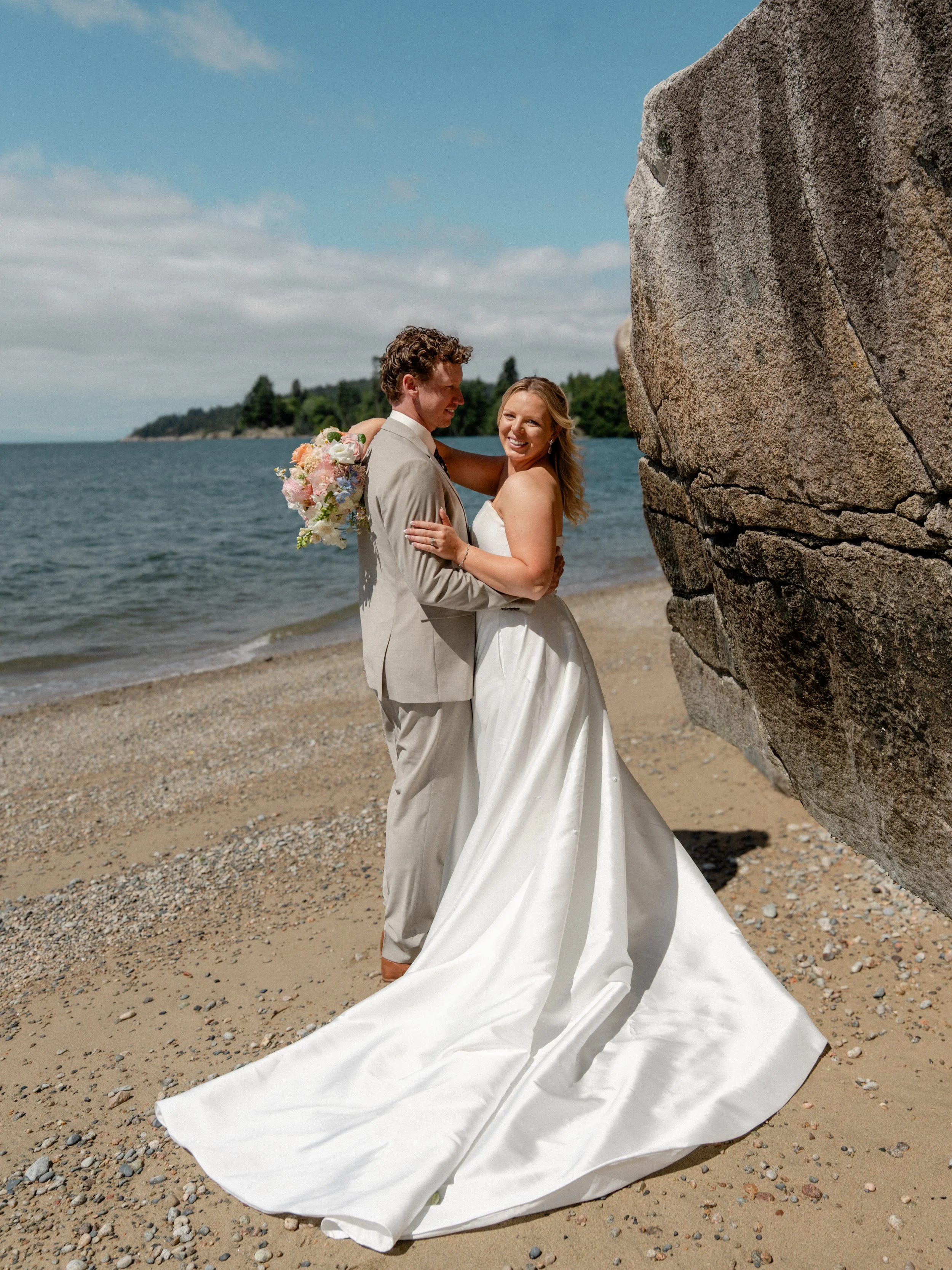 A bride and groom standing on rocks by a lake, with mountains and trees in the background during a cloudy day.