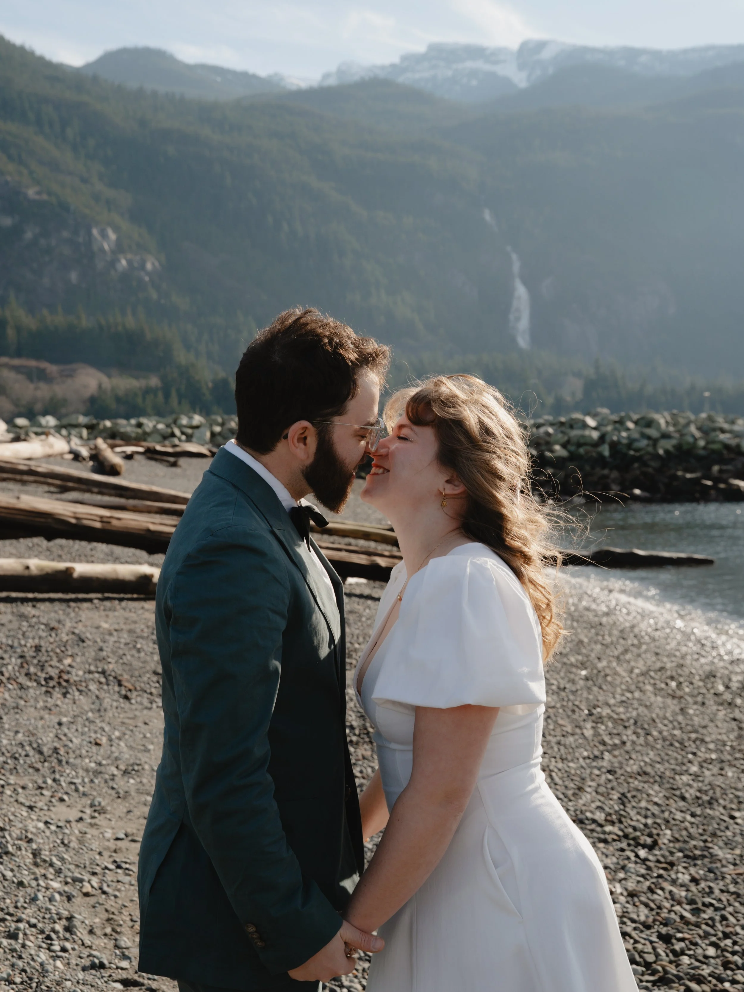 A newlywed couple is standing on a rocky beach in front of a mountain landscape, about to kiss. The groom is wearing a dark suit and the bride is wearing a white wedding dress.