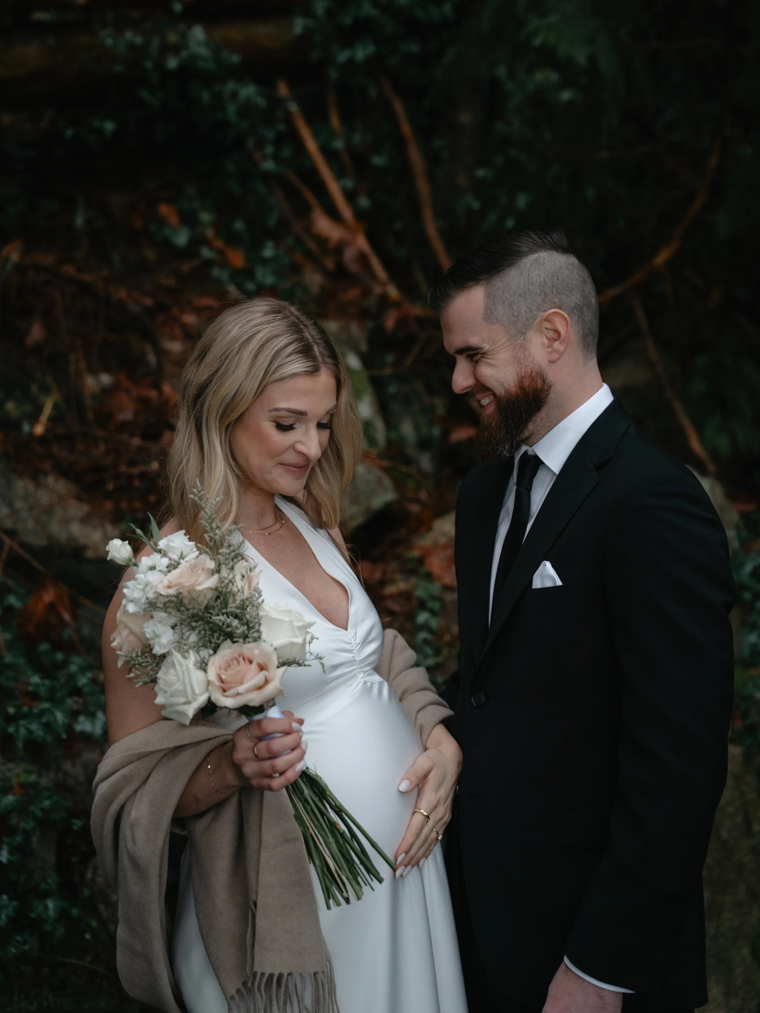 A pregnant woman in a white dress holds a bouquet of pink and white roses while smiling at her partner, a man in a black suit, during an outdoor wedding or photoshoot. They stand against a natural backdrop of greenery.