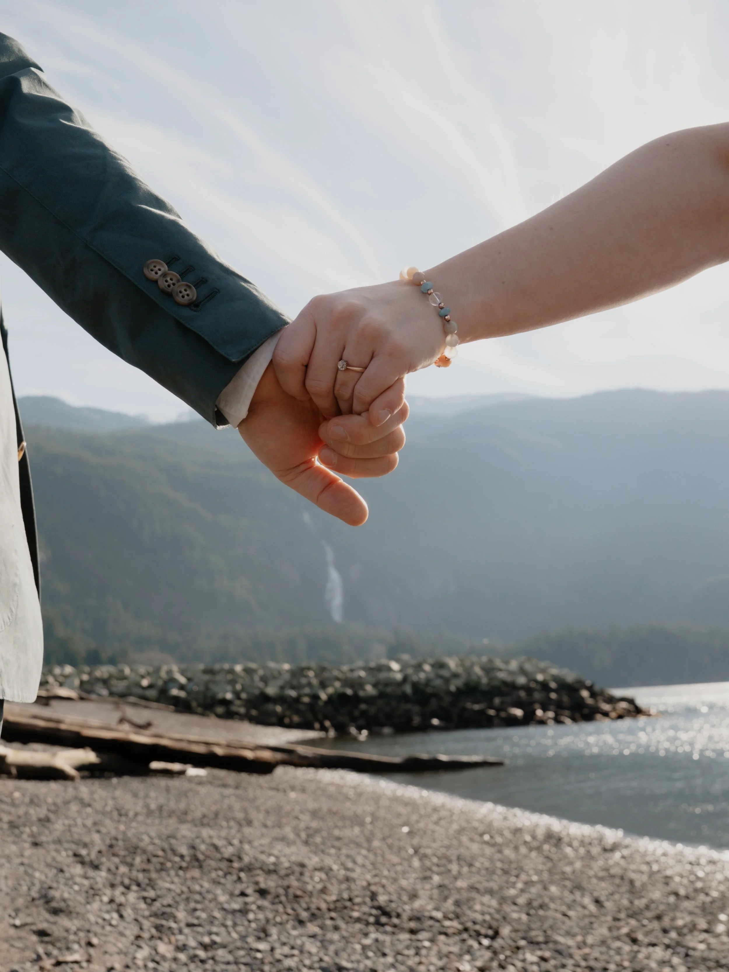 Close-up of a couple holding hands with intertwined fingers, standing on a rocky beach with a body of water and mountains in the background. One person wears a gray jacket and the other wears a bracelet.