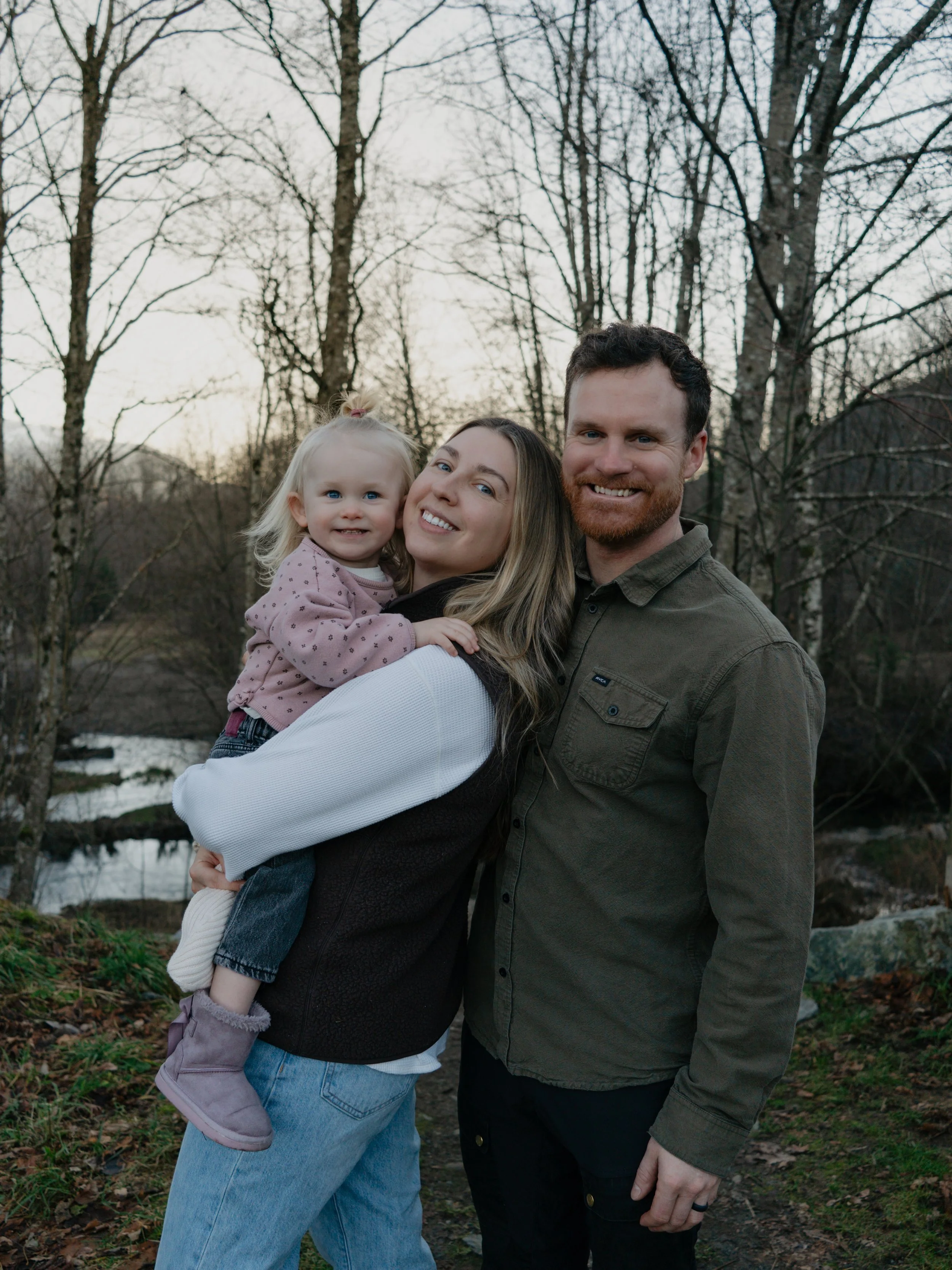 A family of three, a woman, a man, and a young girl, smiling outdoors during sunset with bare trees and a small creek in the background.