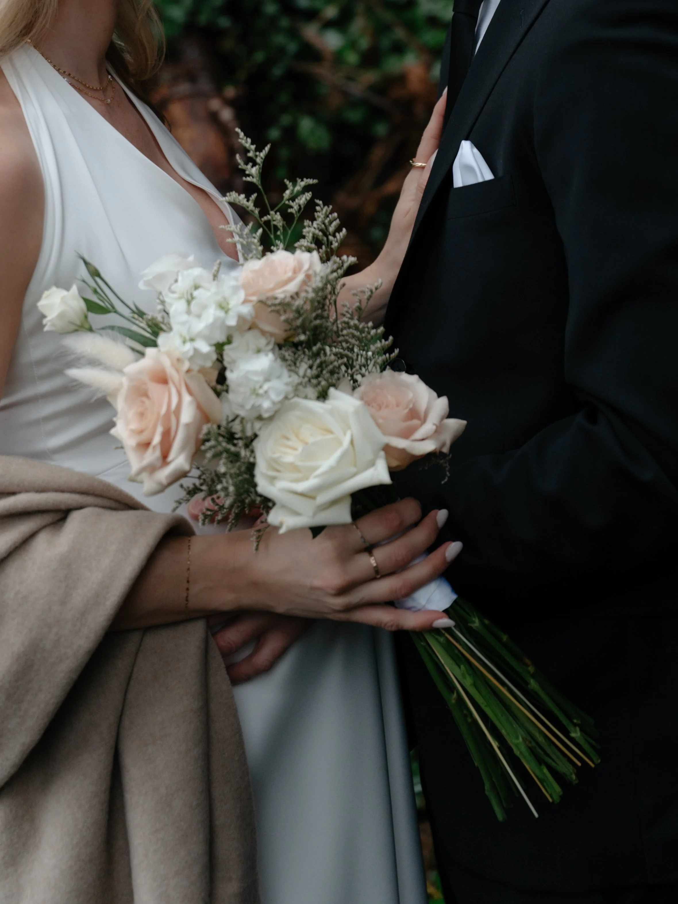 Close-up of a bride holding a bouquet of white and blush pink roses, with a groom dressed in a black tuxedo.