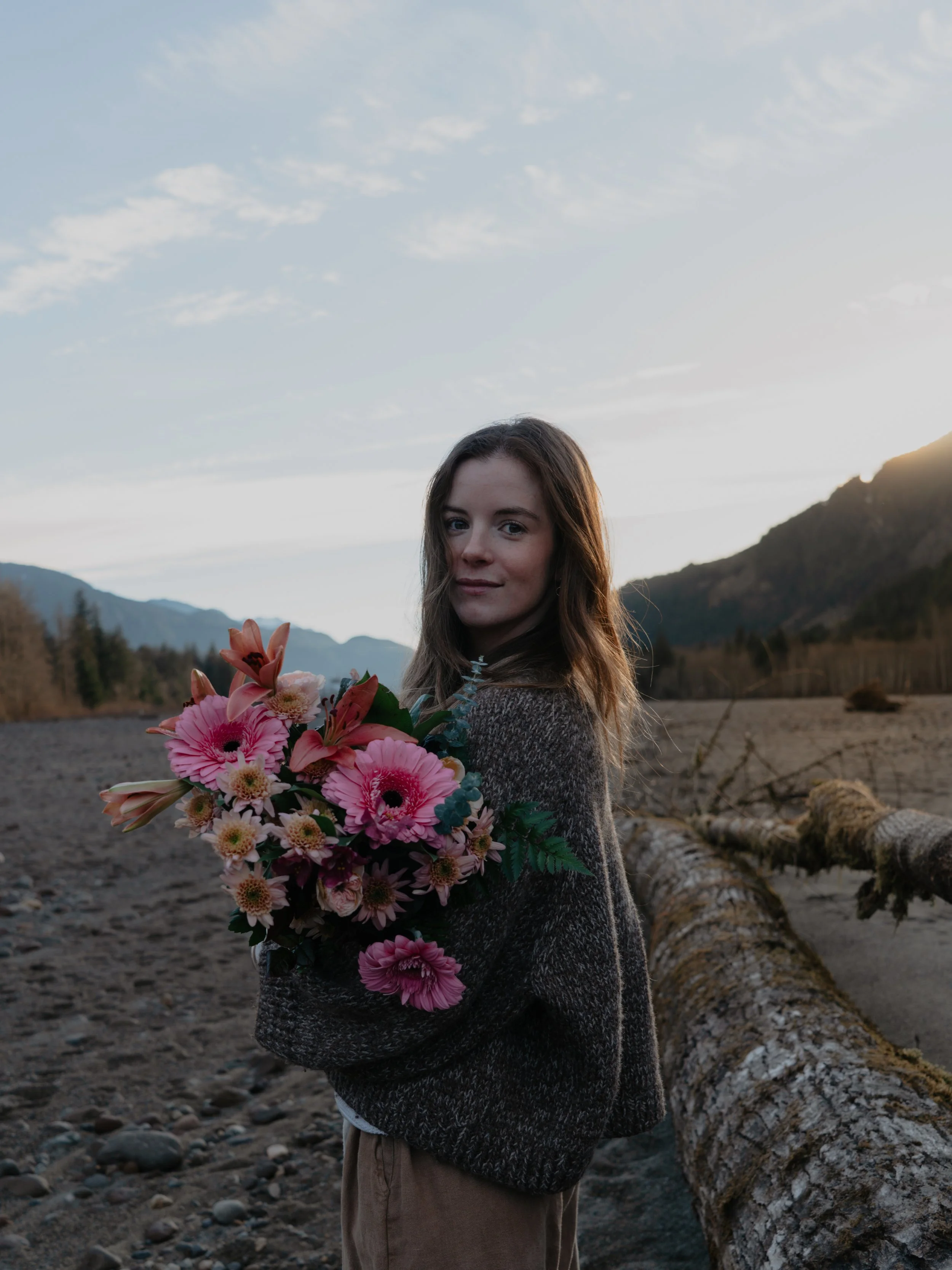 A young woman holding a bouquet of pink flowers standing outdoors in a mountain valley at sunset.