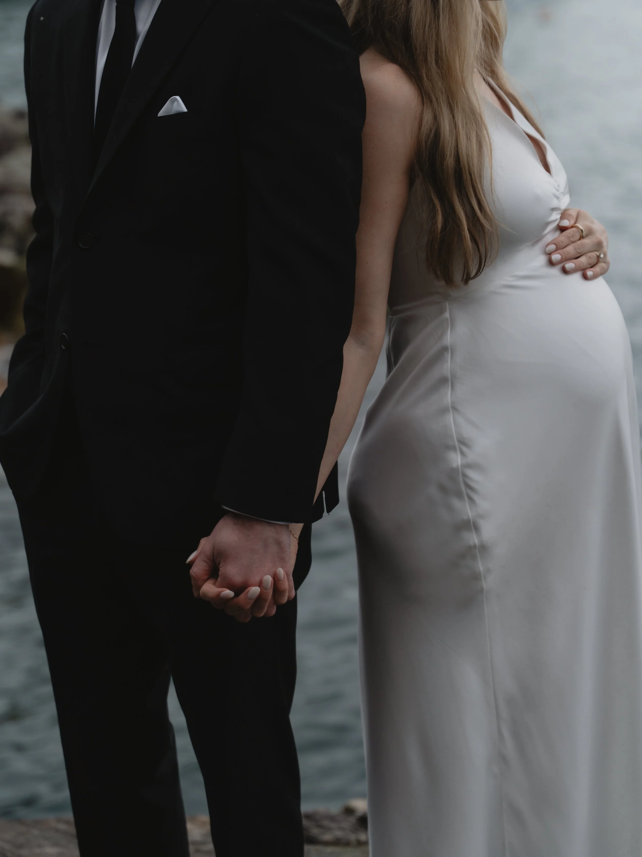A couple holding hands, with a pregnant woman in a white dress and a man in a black suit, standing near a body of water.