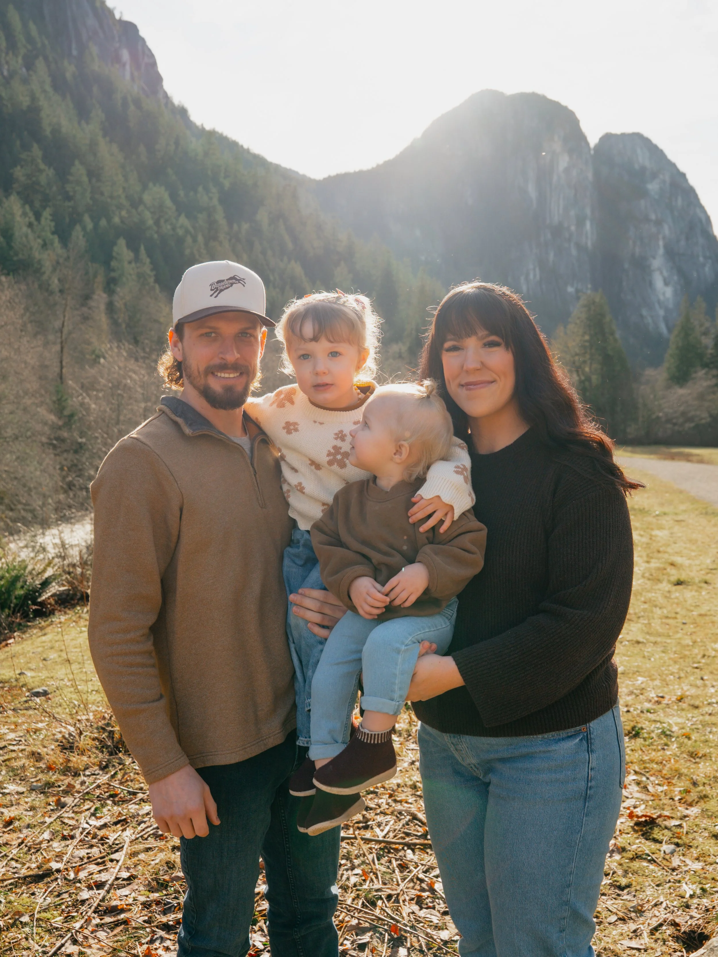 A family of four standing outdoors in front of mountains, smiling and posing for a photo.