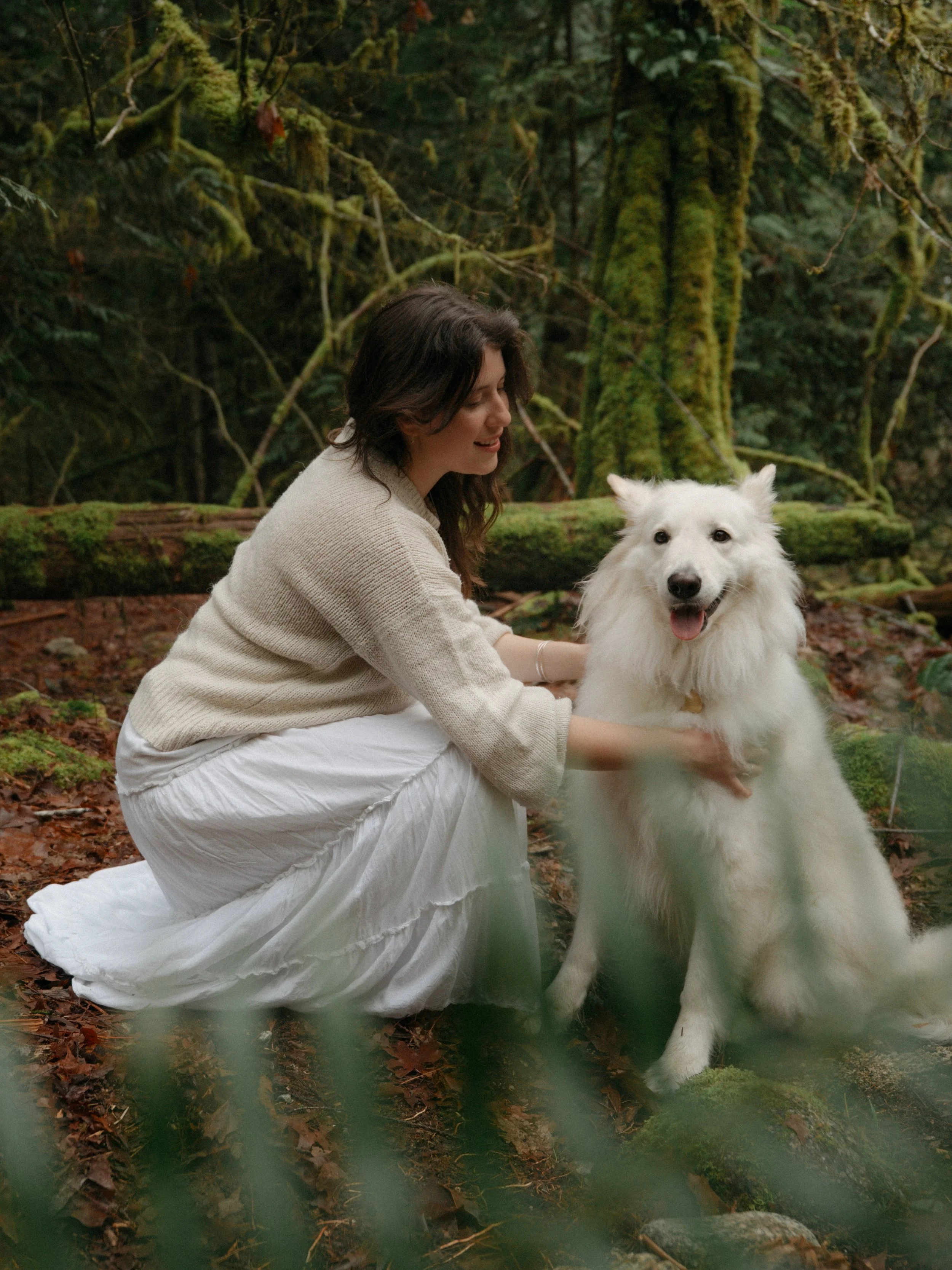 A woman with dark hair wearing a light beige sweater and a long white skirt kneeling on the forest floor, smiling at a large white fluffy dog sitting next to her in a dense green forest.