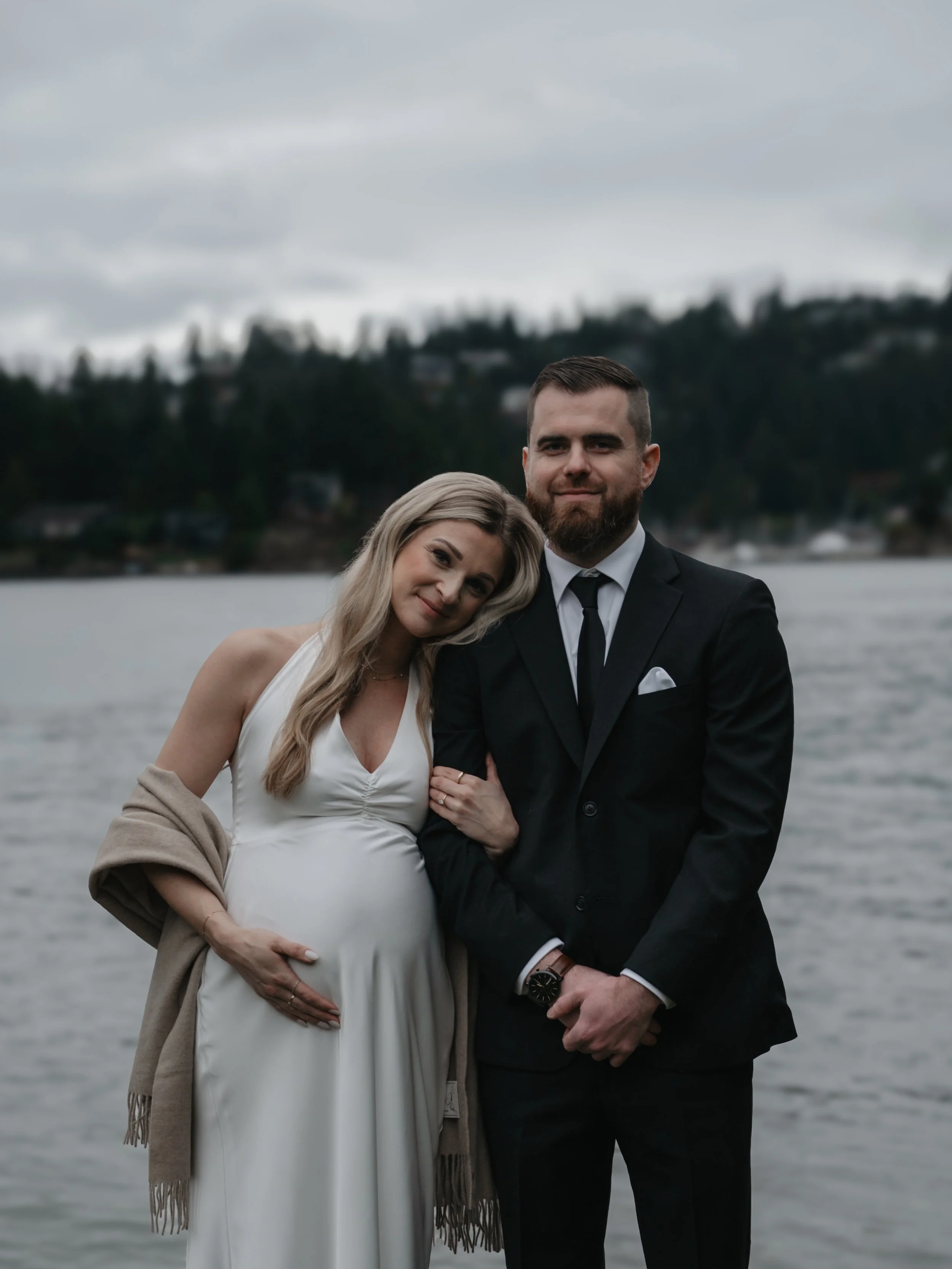 A couple standing by a river with trees and houses in the background; the woman is pregnant and leaning her head on the man's shoulder, both are dressed formally, and it is overcast.