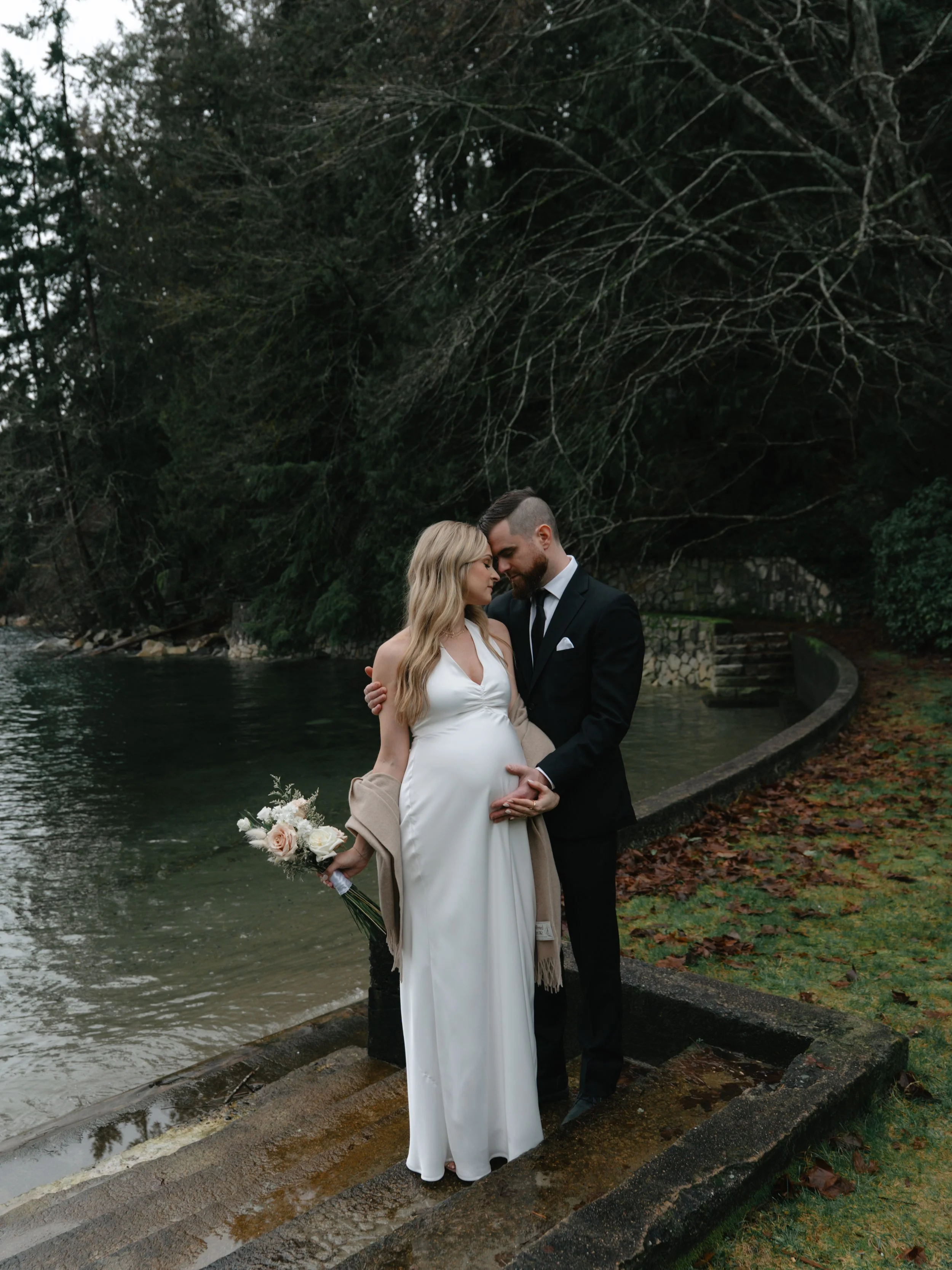 A pregnant woman in a white dress and beige shawl holding a bouquet of flowers, standing close to a bearded man in a black suit and tie, by a river with trees and autumn leaves.
