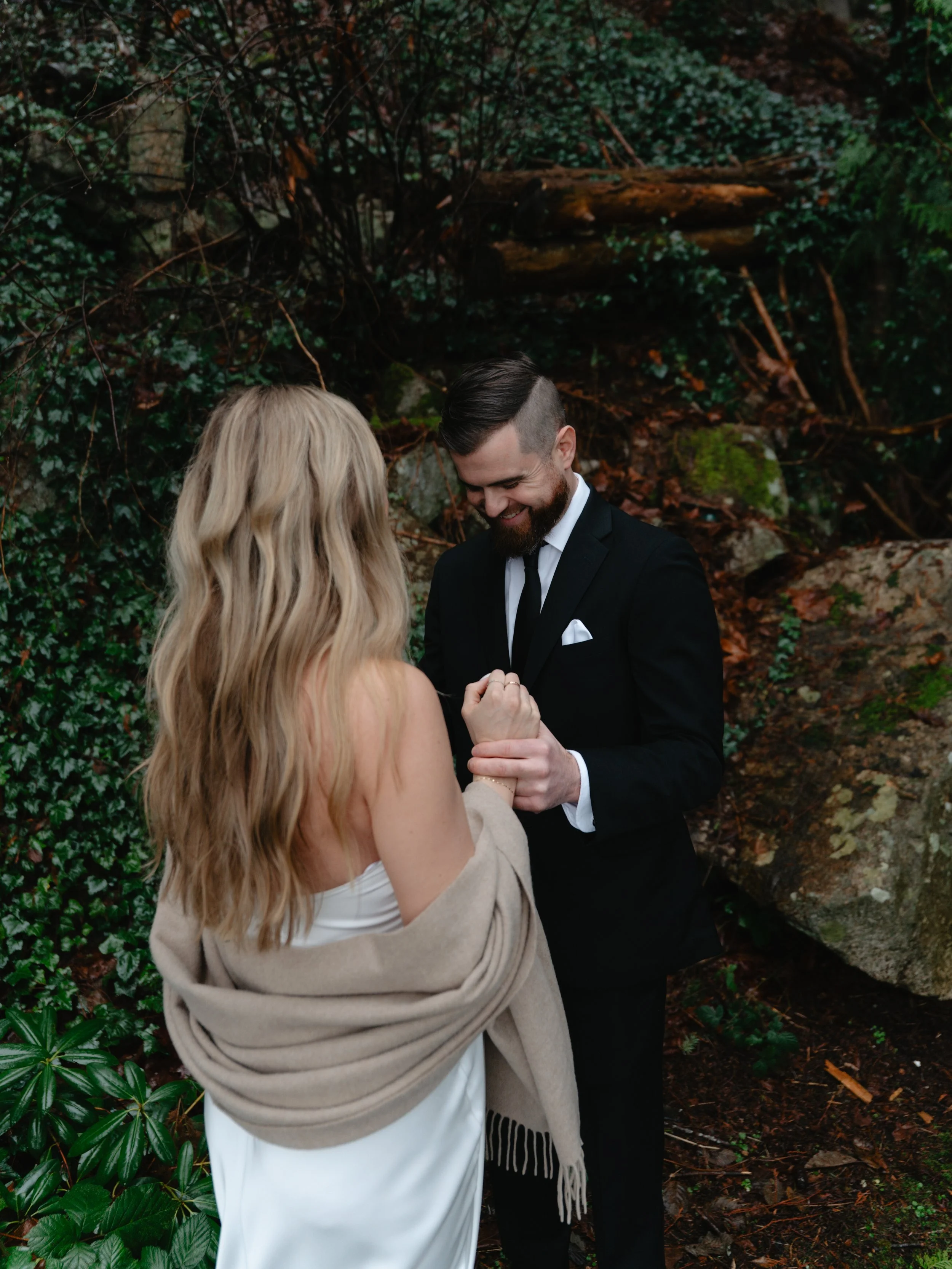 A man in a black suit and tie with a white pocket square is holding the hand of a woman in a strapless white dress and a beige shawl, with an outdoor woodland backdrop of rocks and dense greenery.