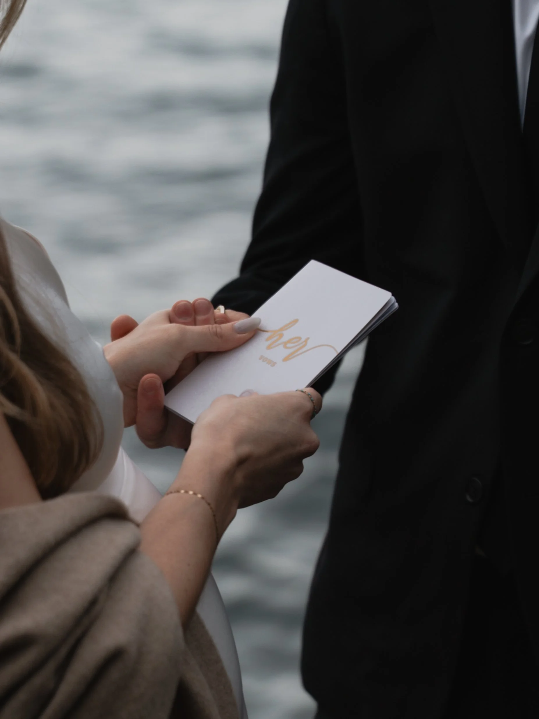 Close-up of a woman holding a small white card with gold cursive writing, partly reading 'her,' during a wedding ceremony by the water.