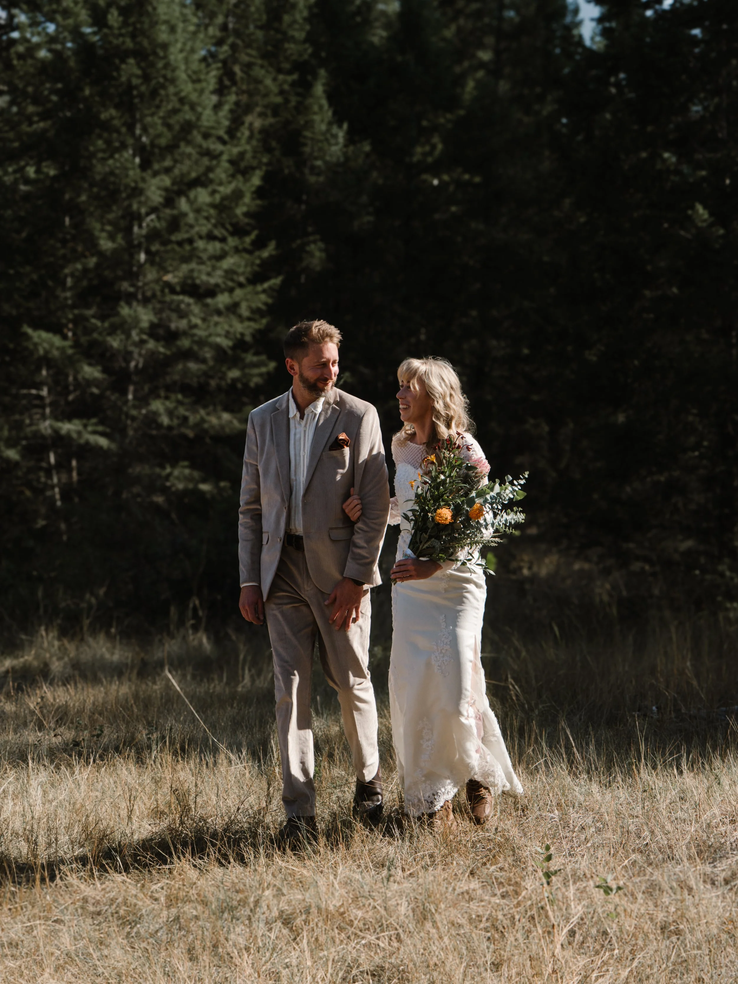 A couple walking outdoors in a grassy area, with the woman holding a bouquet of flowers. The woman is wearing a white wedding dress and the man is dressed in a light-colored suit. They are smiling and looking at each other, surrounded by trees.
