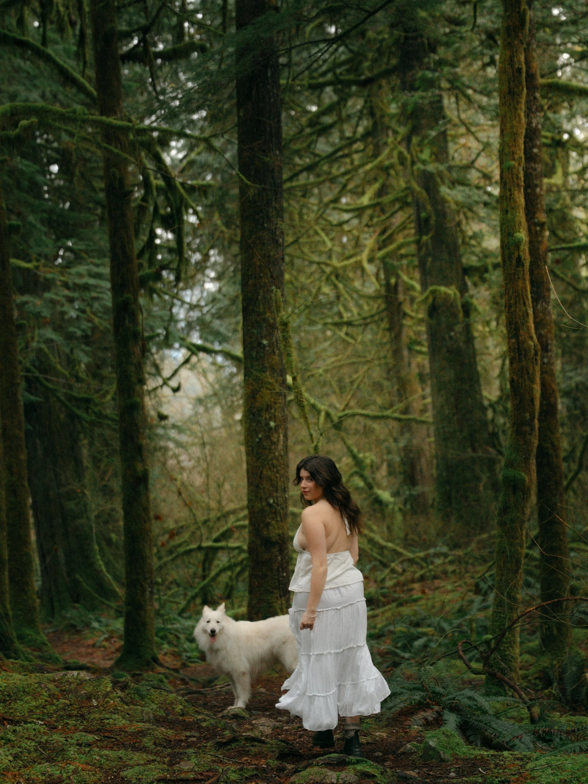 A woman in a white dress standing on a forest trail with a white fluffy dog nearby, surrounded by tall moss-covered trees.