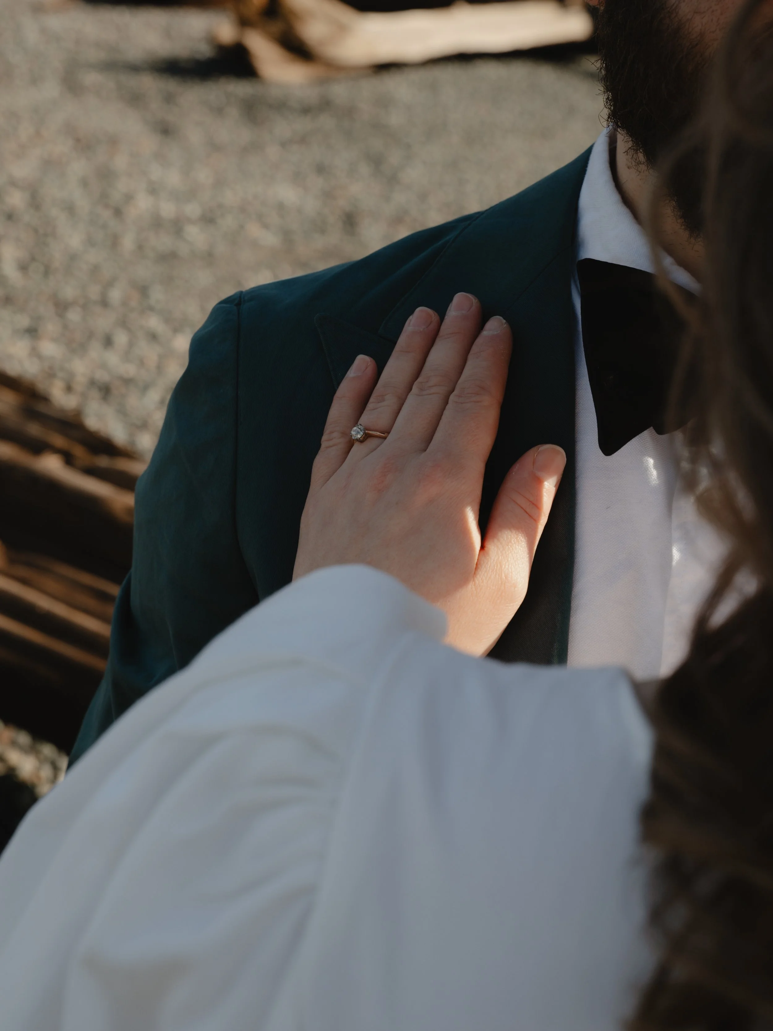 Close-up of a woman's hand resting on a man's shoulder, displaying an engagement ring with a diamond. The man is dressed in a tuxedo with a bow tie, and the background appears to be a wooden structure outdoors.