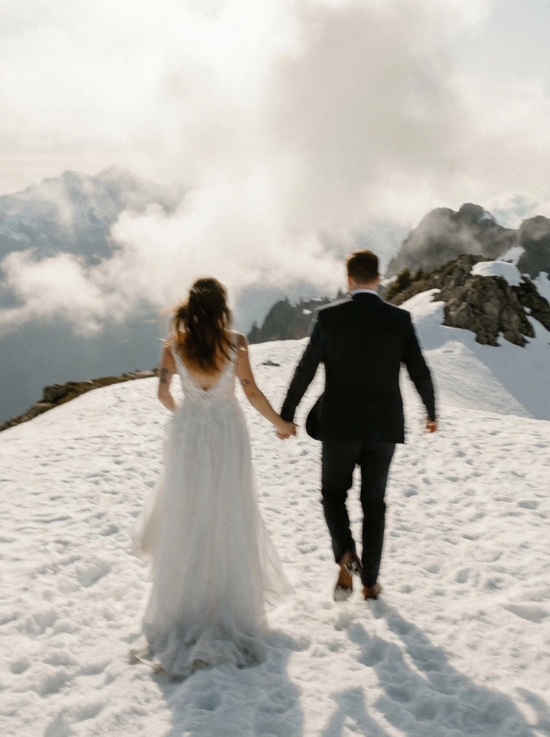 A bride and groom walking hand in hand on snowy mountain terrain with cloudy sky and mountain peaks in the background.