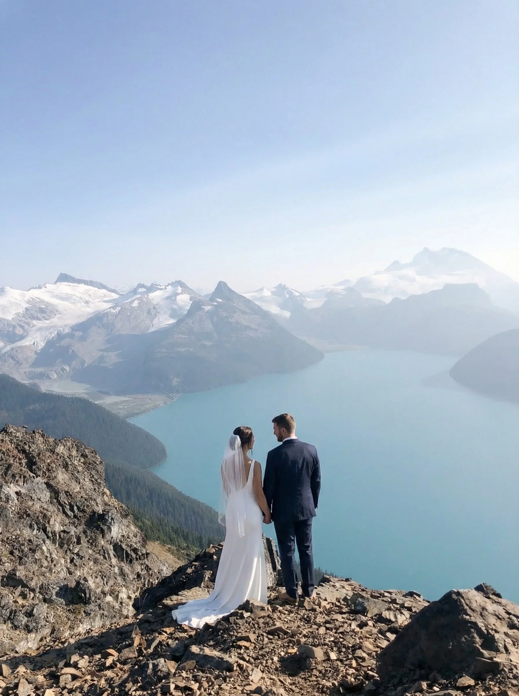 A bride and groom standing on rocky terrain overlooking a mountain lake with snow-capped peaks in the background.