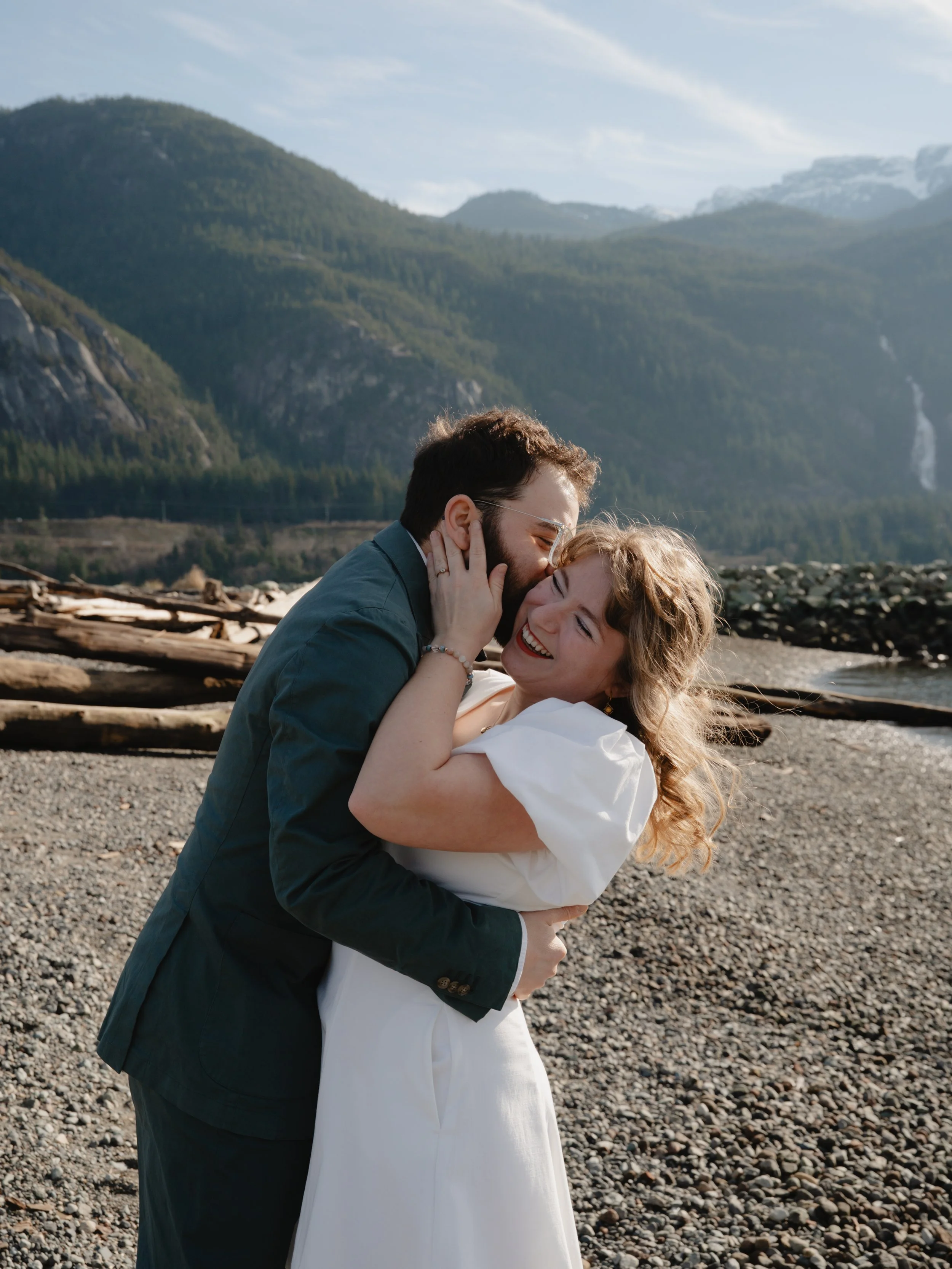 A couple dressed in formal attire, embracing and sharing a joyful moment on a rocky beach with mountains in the background.