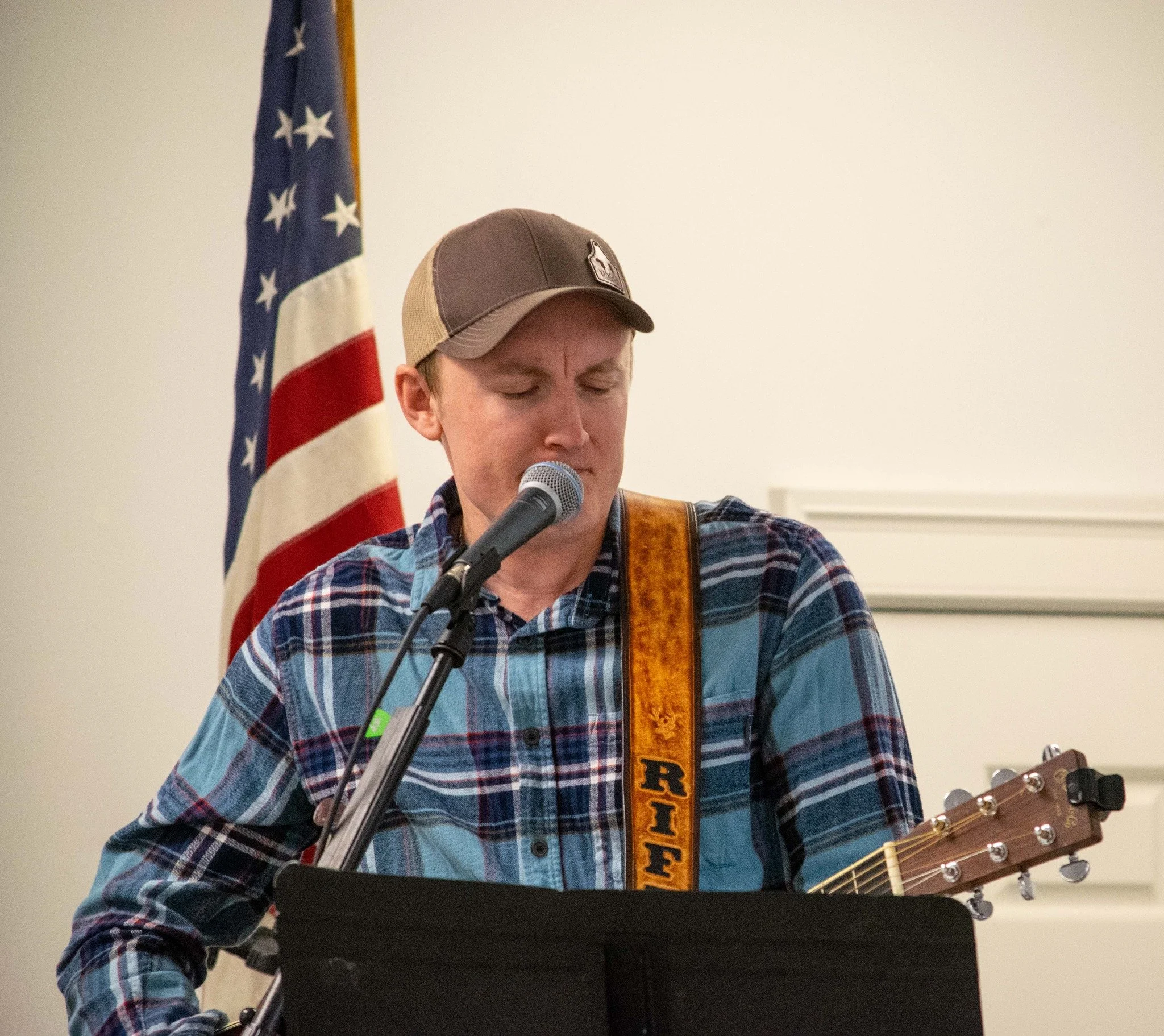 Young man with a plaid shirt singing into a microphone while playing a guitar, with an American flag in the background.