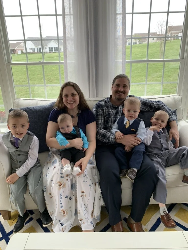 Family of seven sitting on a white couch in a living room with large window showing green lawns and houses outside.