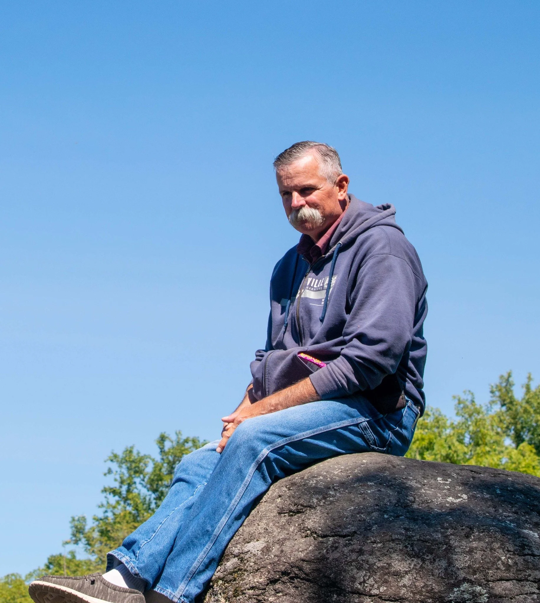 A man with a gray mustache and gray hair, wearing a dark hoodie and jeans, sitting on a large rock outdoors against a blue sky background.