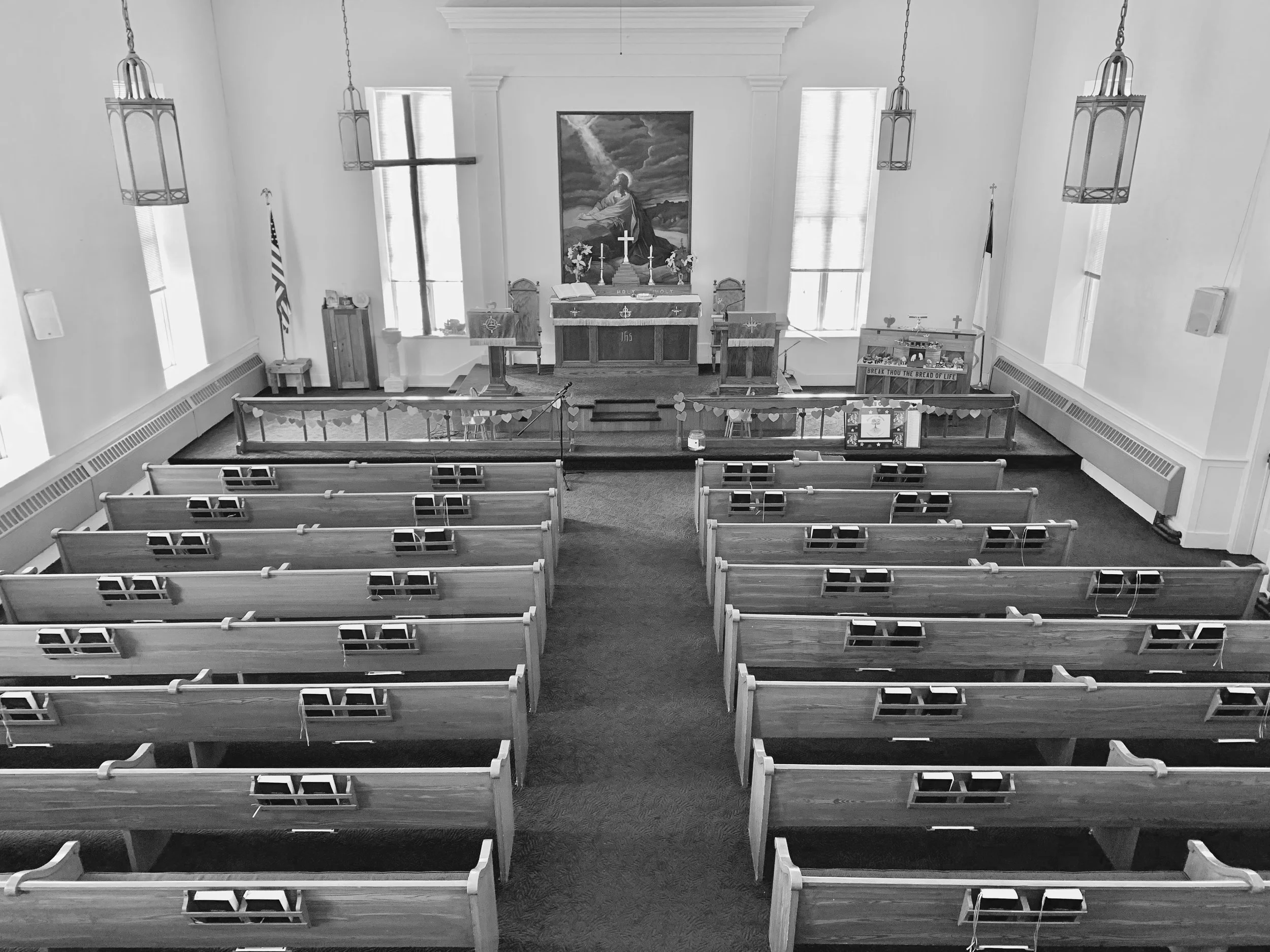 A black and white photo of the interior of a church with pews facing an altar. The altar has three crosses, an open book, and candles. Behind it is a large painting of Jesus. There are windows on either side of the altar and hanging light fixtures from the ceiling.