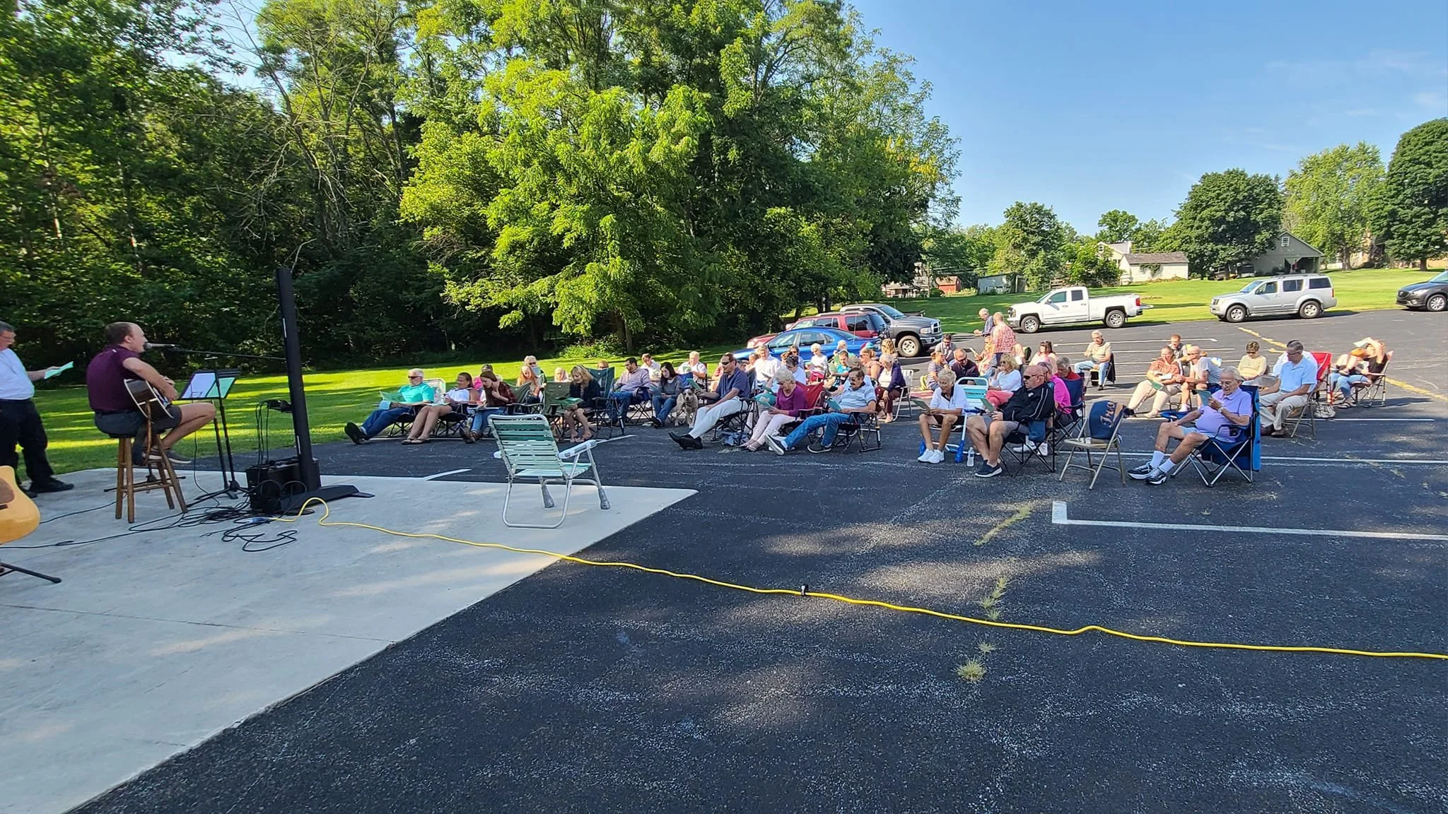 Outdoor gathering with people sitting in chairs watching a live music performance on a small stage, with trees and parked cars in the background on a sunny day.