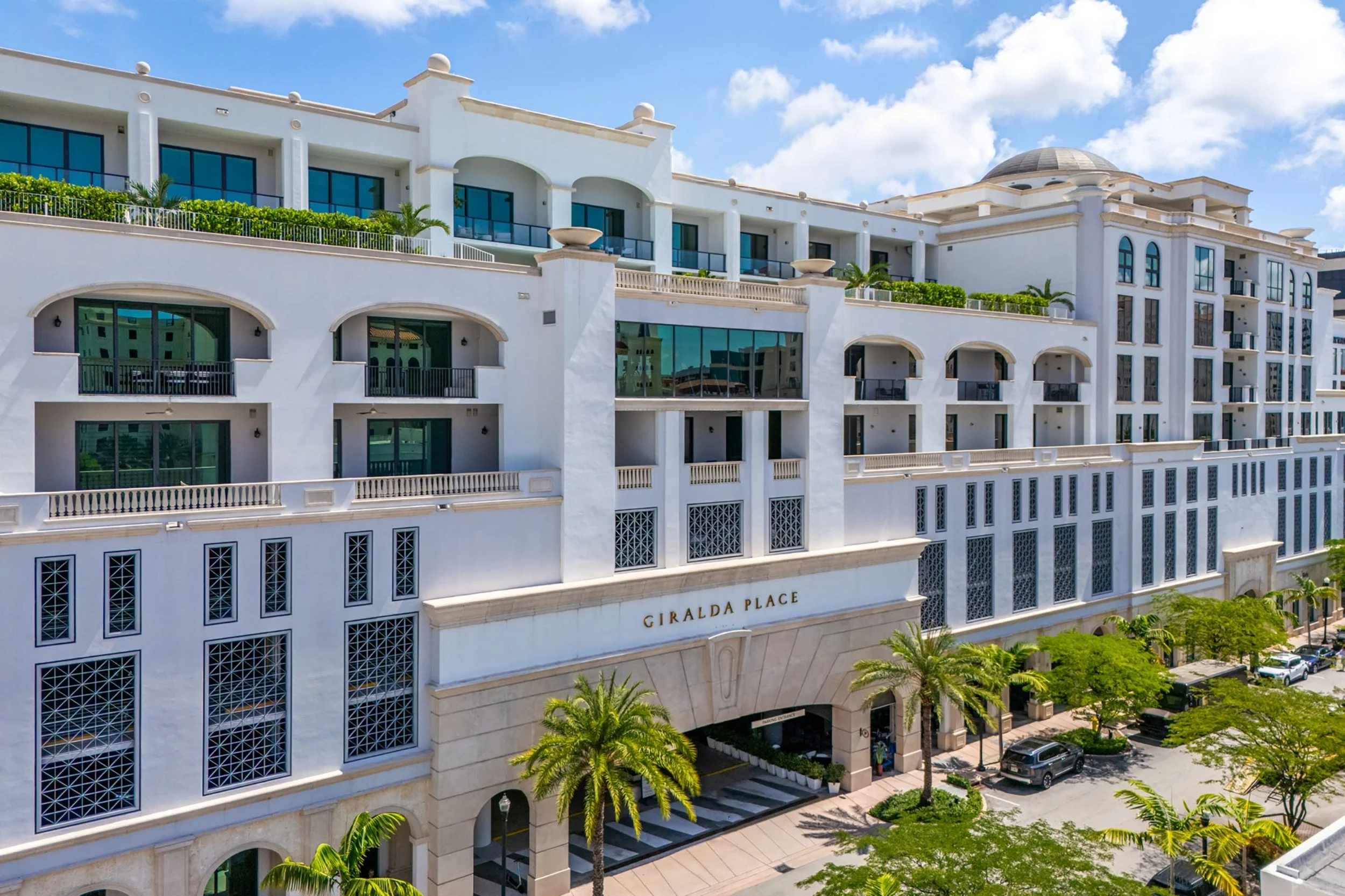 Front view of a modern white hotel named Giralda Place with multiple balconies, palm trees, parked cars, and a bright blue sky with clouds.