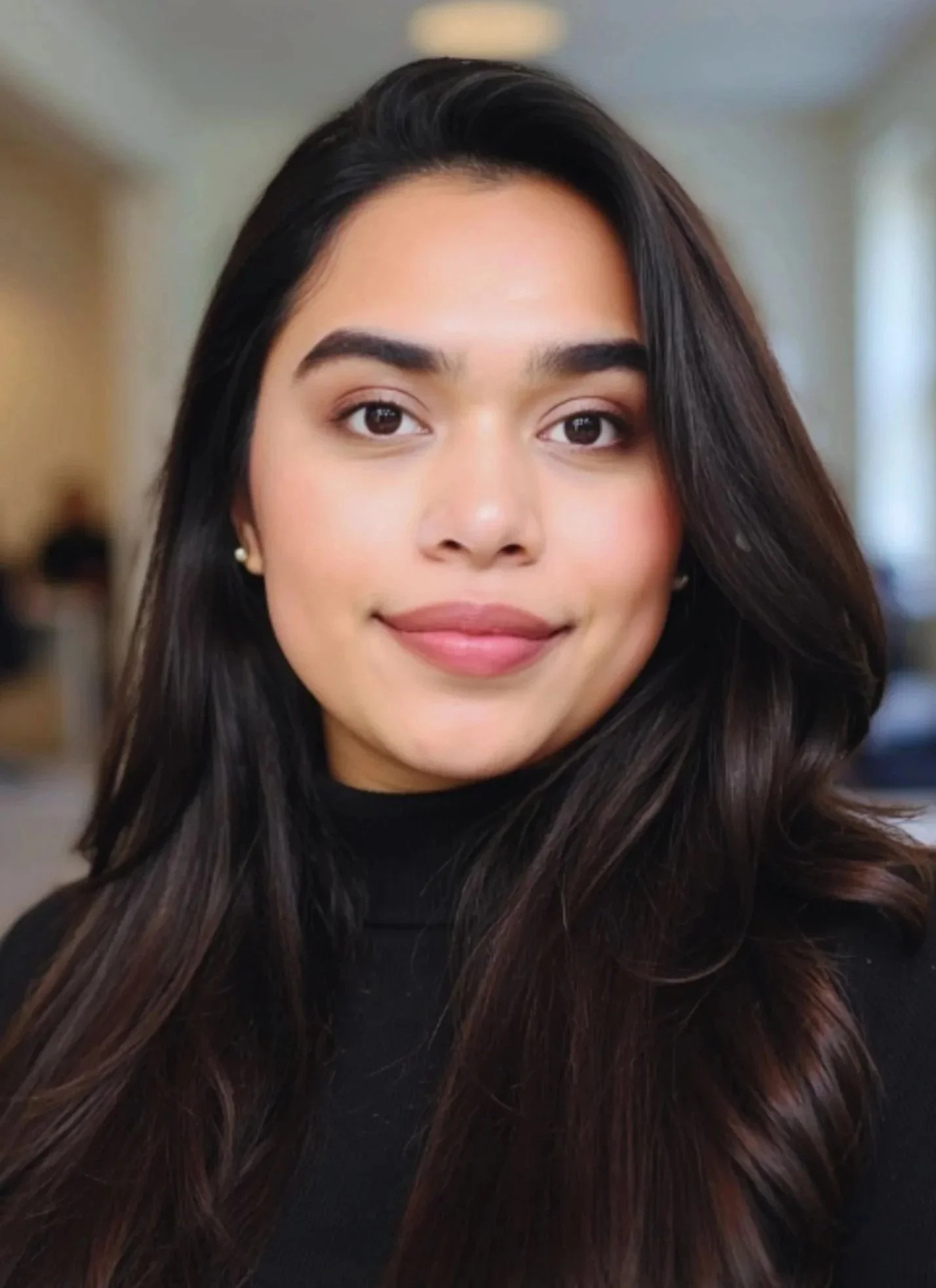 Close-up of a young woman with long, dark hair wearing a black top, smiling softly, in a softly lit indoor setting.